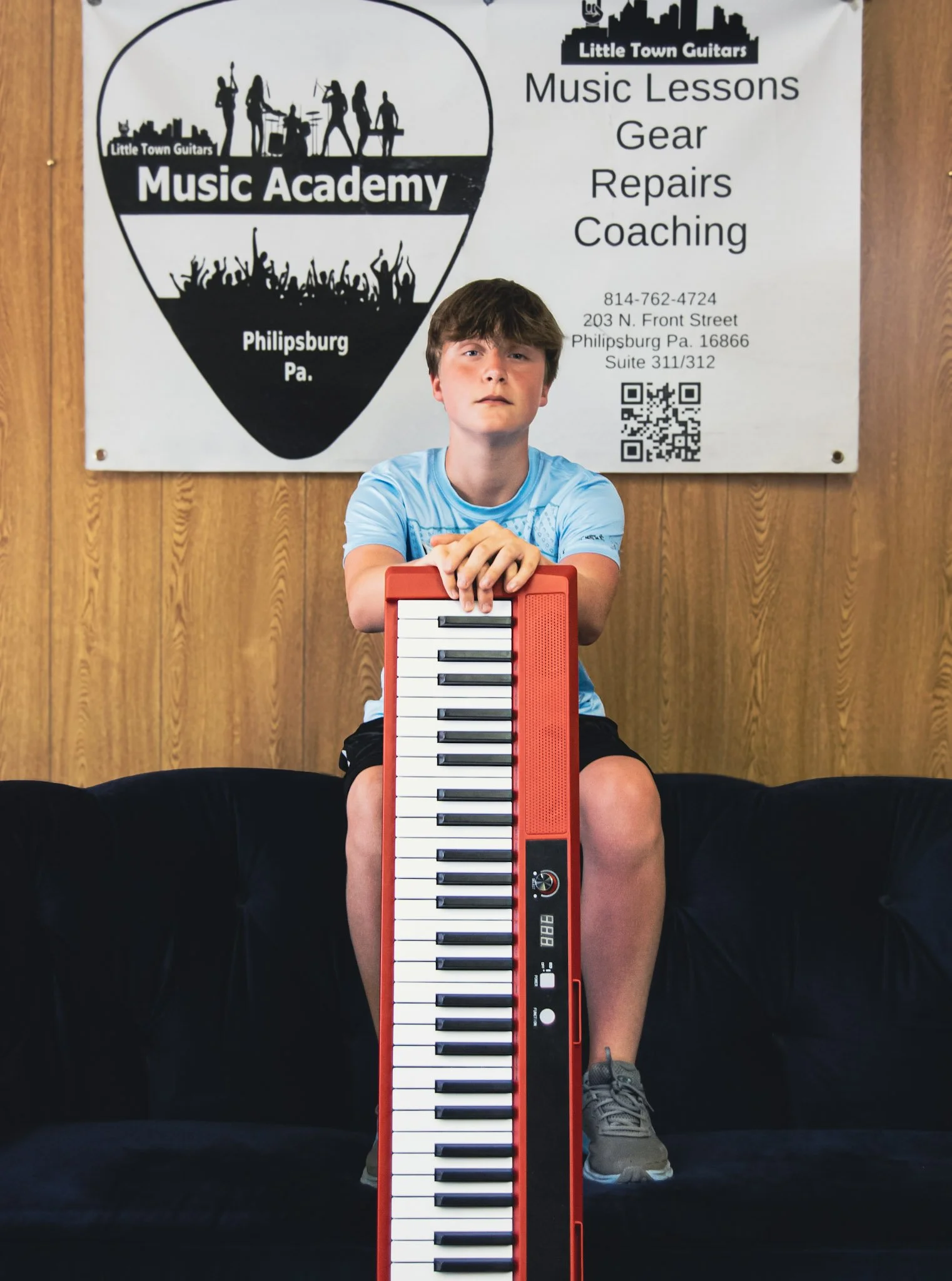 A young boy sitting on a black couch holding a red keyboard piano in front of him. There is a large sign behind him with information about music lessons, gear, repairs, and coaching at Little Town Guitars in Philipsburg, PA.