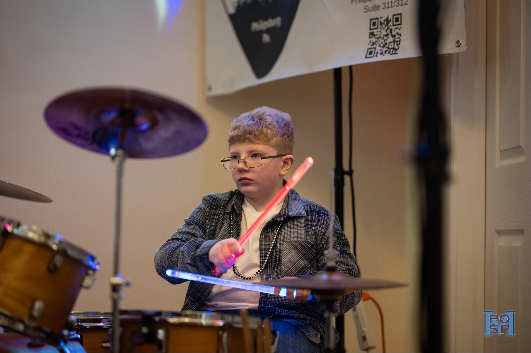 A young boy with glasses playing drums with a pink LED drumstick, in a room with a beige wall and a poster in the background.