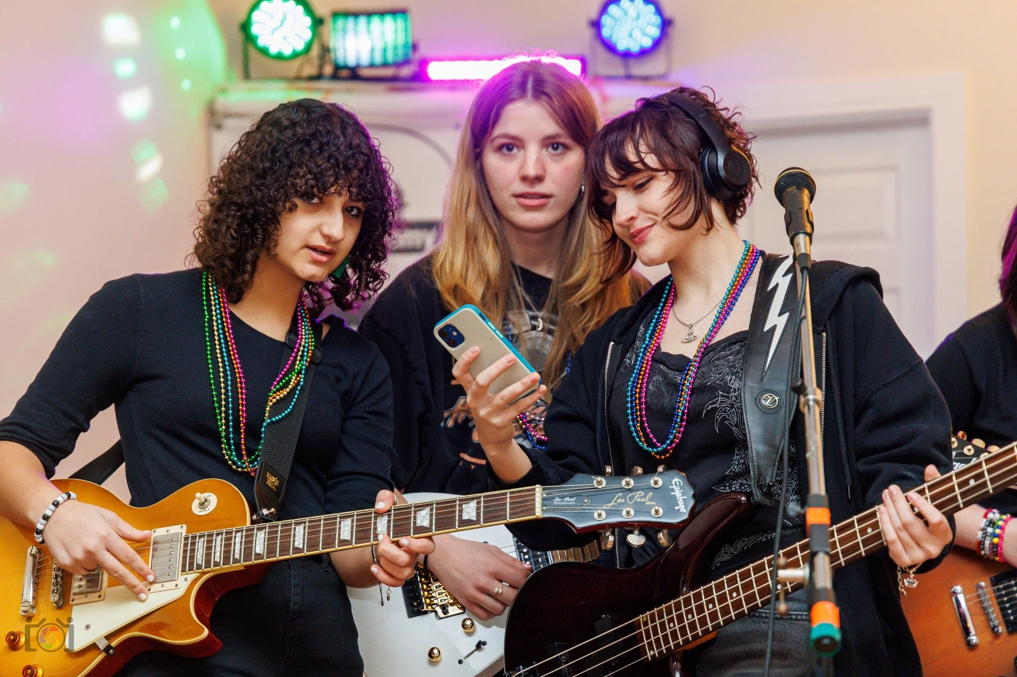Three young women in a band practicing, with two playing guitars and one holding a smartphone, wearing colorful bead necklaces and casual clothing.
