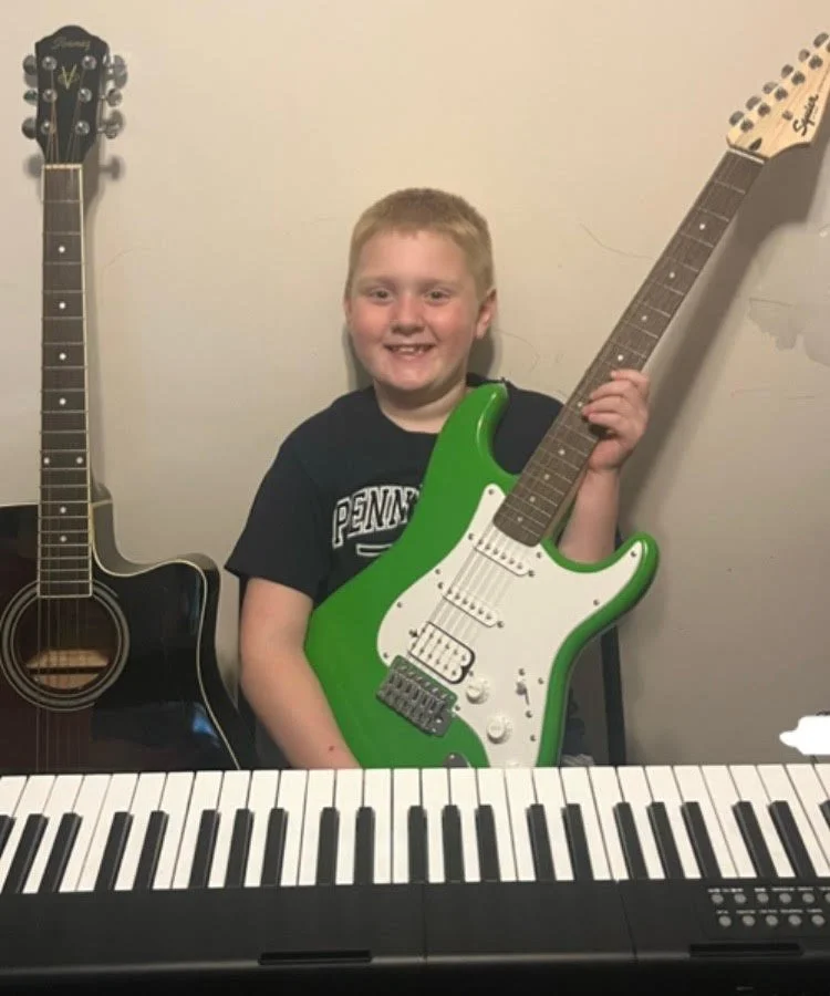 A boy holding a green electric guitar, sitting in front of a keyboard, with two acoustic guitars on either side.