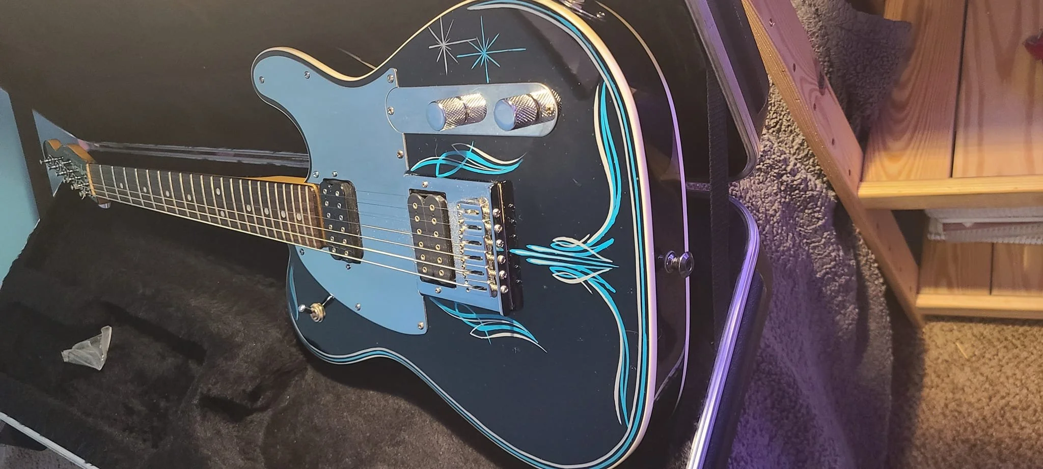 Black electric guitar with blue and silver flame design, chrome hardware, and a glossy finish, resting on a black case in a room with wooden furniture and carpeted floor.