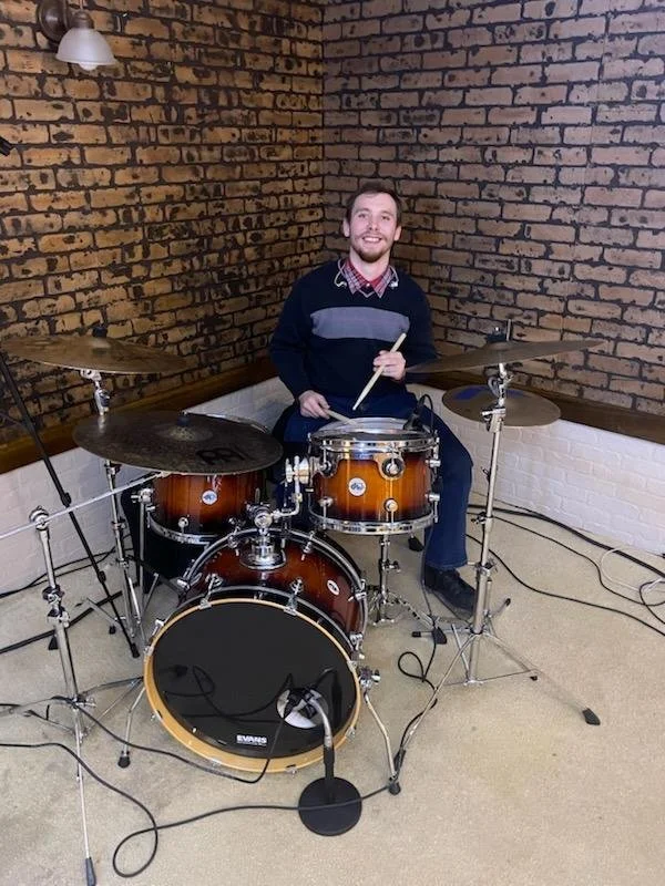 A man sitting behind a drum set in a room with exposed brick walls, smiling at the camera.