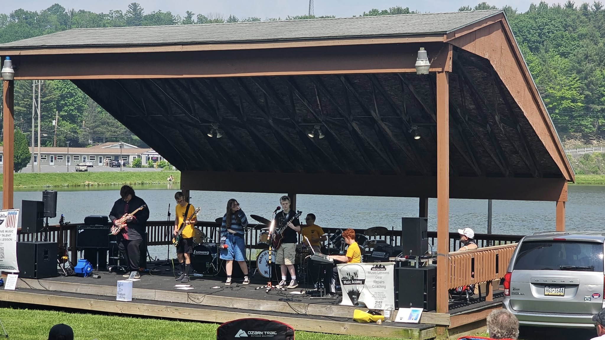 A band performing on an outdoor stage located by a lake, with a water and trees in the background. The band members are playing instruments including guitars, drums, and keyboard. There is a large gray vehicle parked nearby and an audience watching.