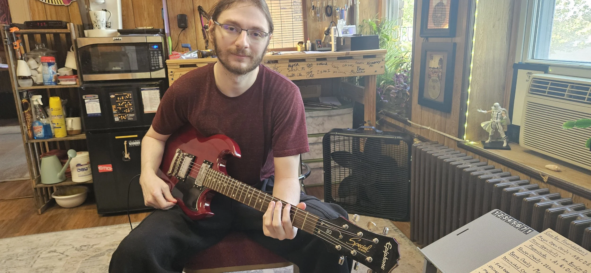 A man with glasses and a beard sits on a stool holding a red electric guitar in a cozy, cluttered room with wooden walls. The room has a microwave, shelves with various items, a window with a small figurine, and a radiator.