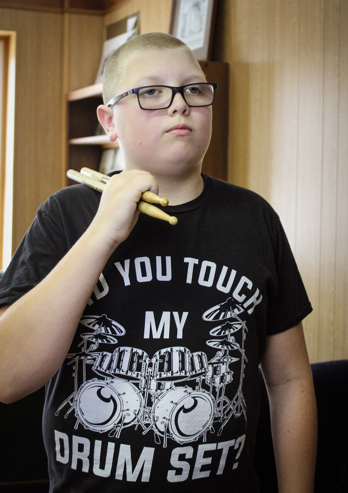 A young boy with glasses holding drumsticks over his shoulder, wearing a black T-shirt with a drum set graphic and the text "Did you touch my drum set?" standing indoors with wood paneling in the background.