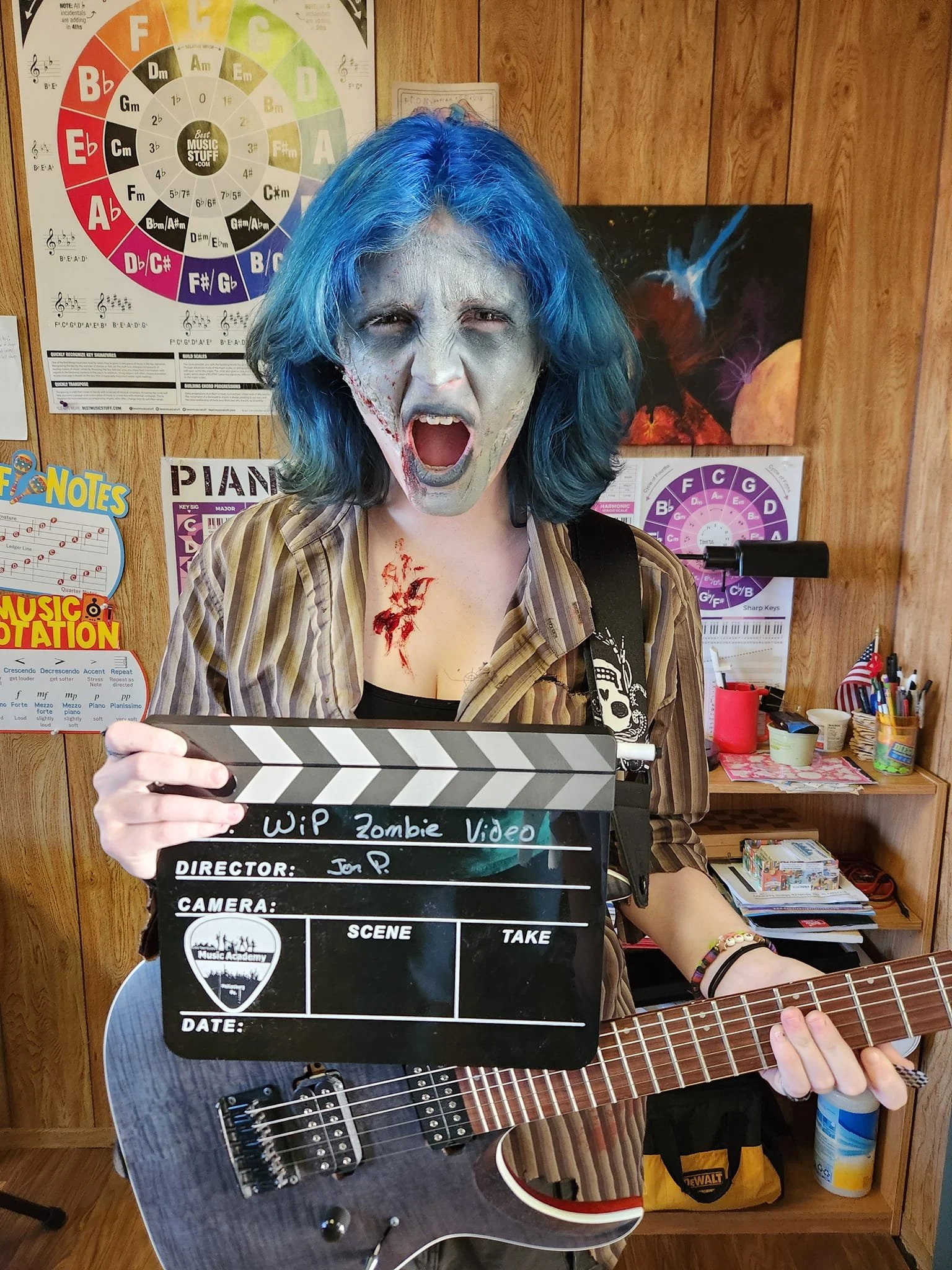 Person dressed as a zombie holding a film slate and playing an electric guitar in a classroom with musical posters and wood-paneled walls.