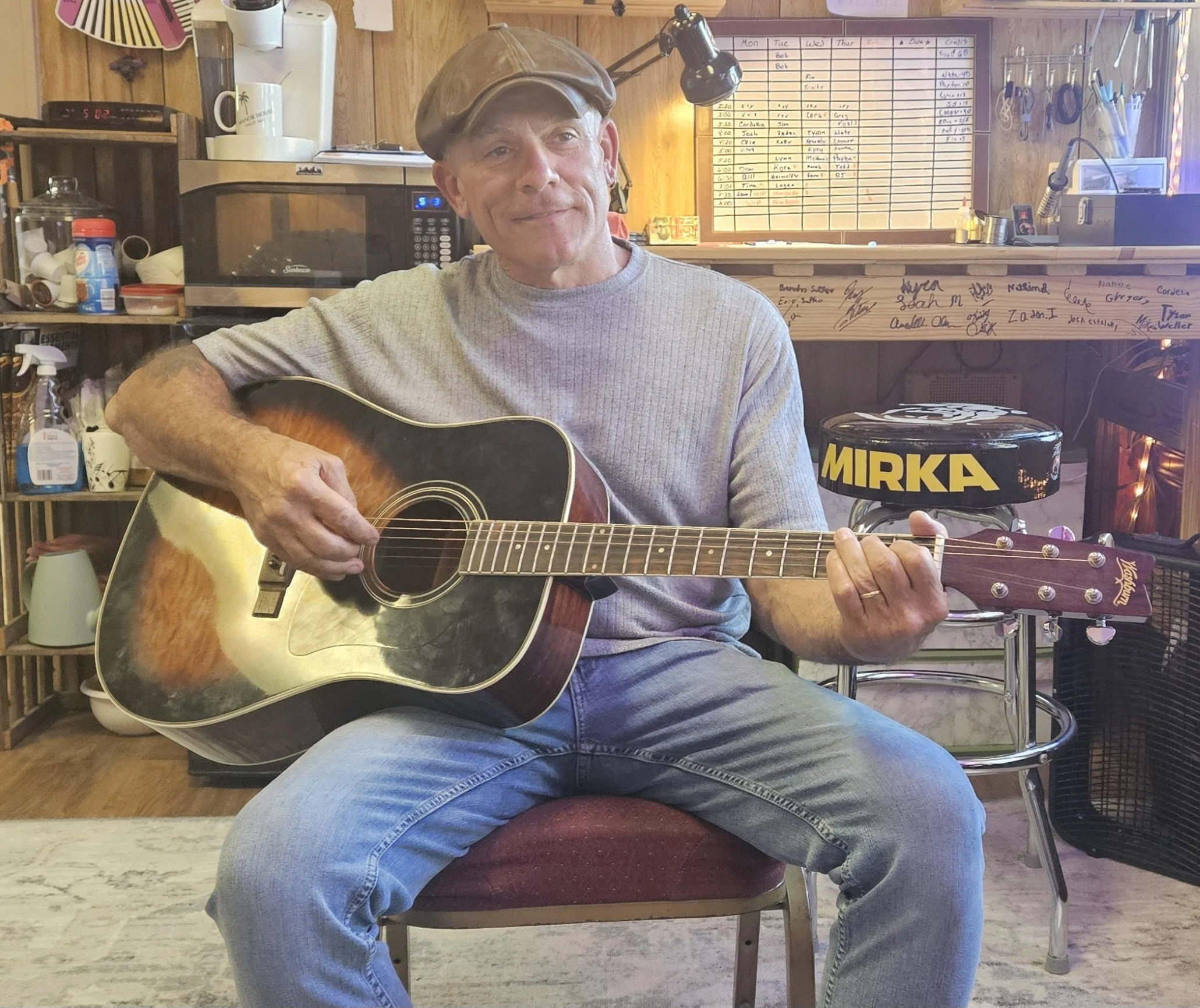 A man in a gray t-shirt and jeans playing an acoustic guitar, seated in a room with a wooden background. The man is wearing a brown cap and smiling. Behind him are shelves with various items and a wall with a large calendar or schedule.