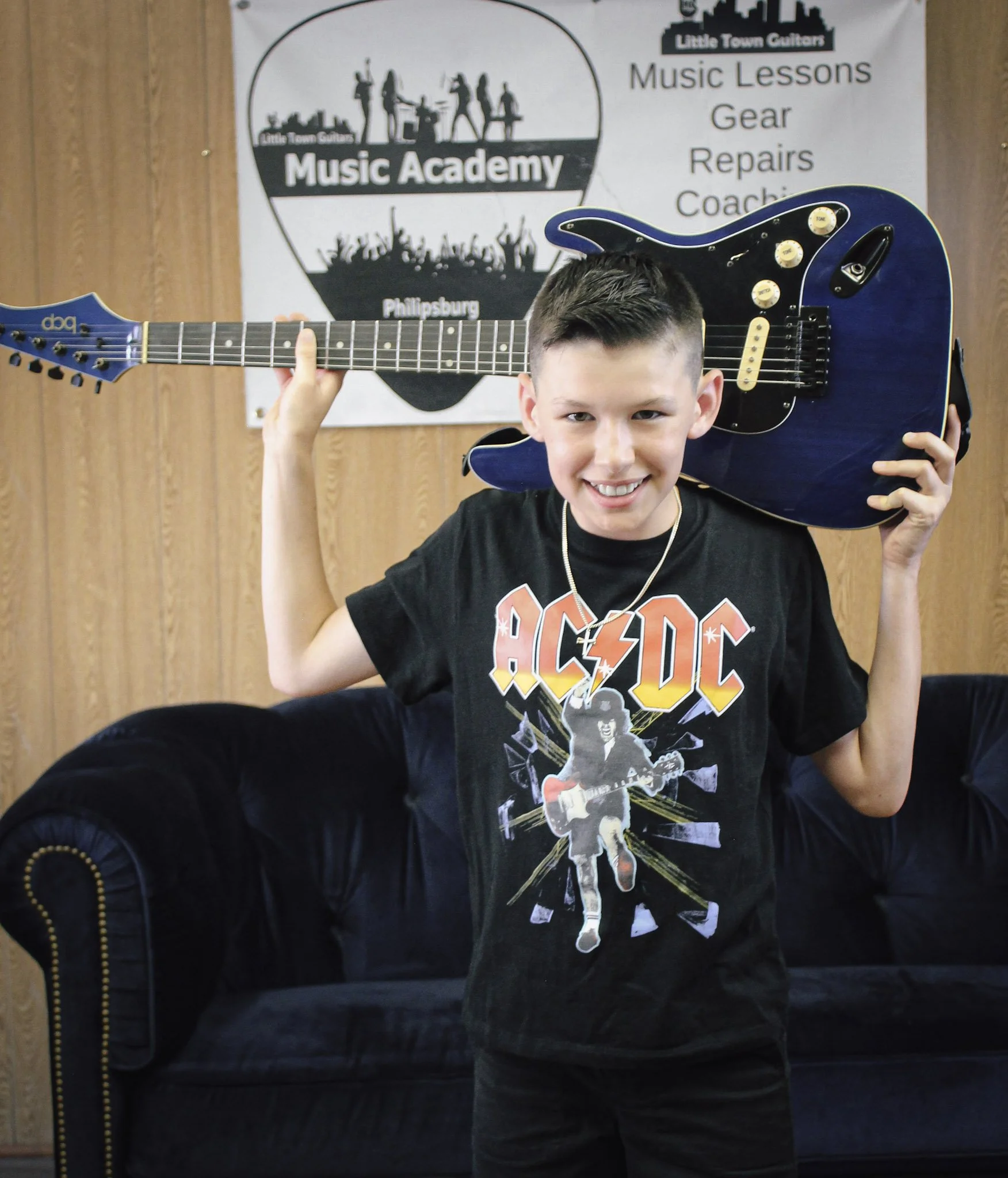 A young boy smiling and holding a blue electric guitar on his shoulder inside a music school. The background features a sign for Little Town Guitars Music Academy with words like 'Music Lessons', 'Gear', 'Repairs', and 'Coaching'.