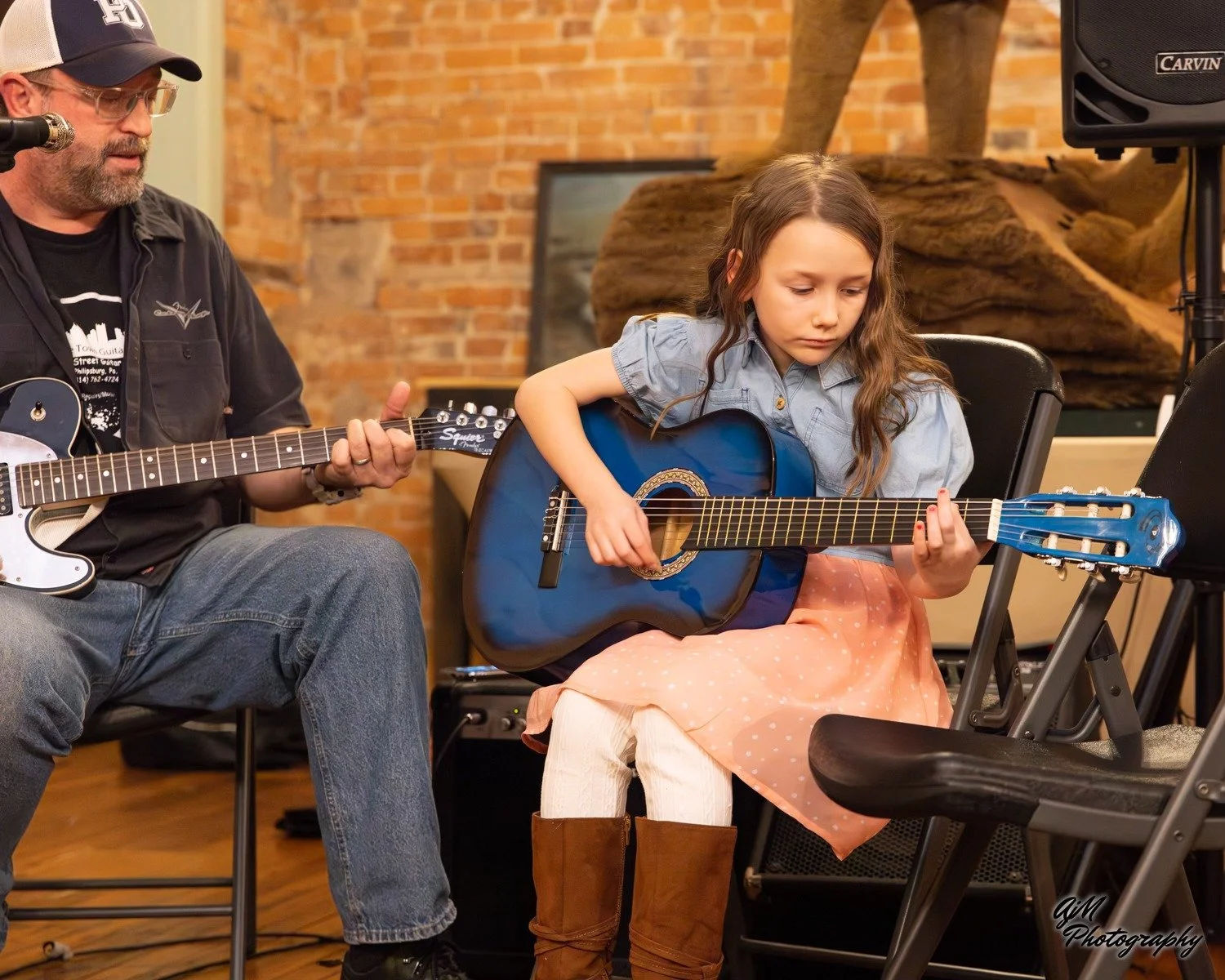 A young girl playing a blue acoustic guitar while sitting on a chair, with an older man playing a black and white electric guitar next to her, in a room with brick walls and wooden accents.