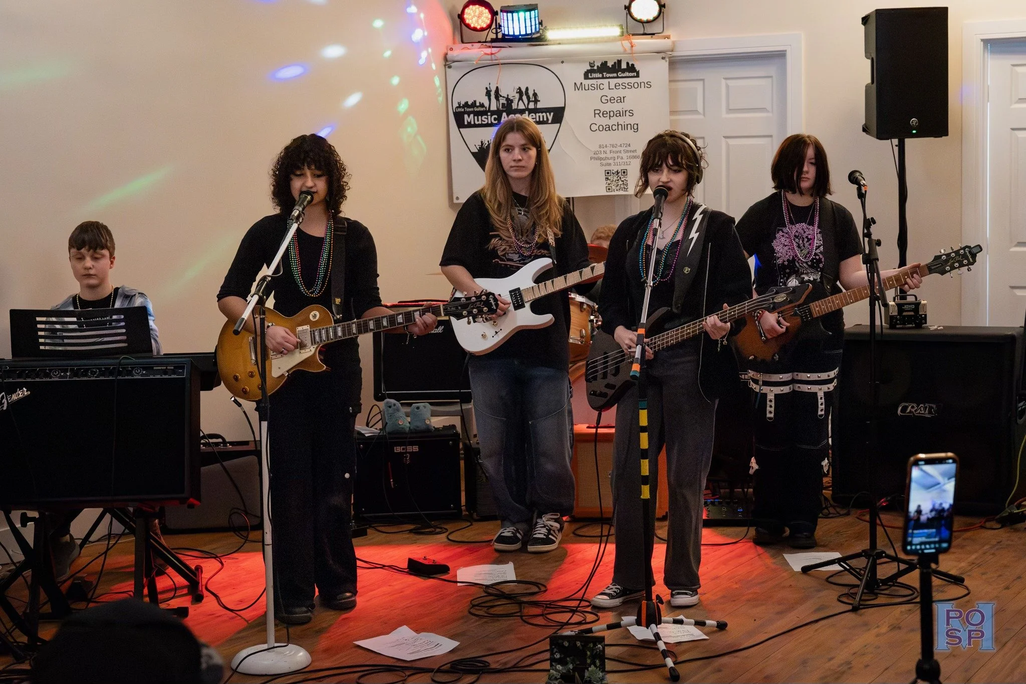 Group of five kids performing with guitars and microphone in a music practice room with a banner in the background that says 'Music Lessons, Gear, Repairs, Coaching'.