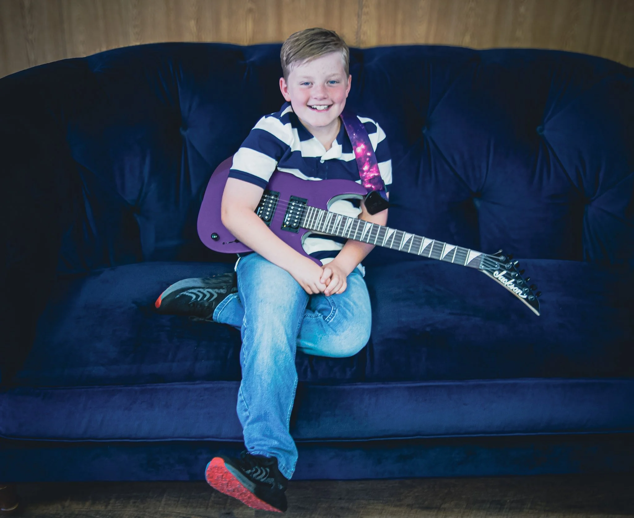 A young boy sitting on a dark blue velvet sofa, holding a purple electric guitar, wearing a black and white striped shirt, blue jeans, and black sneakers with red soles, smiling at the camera.