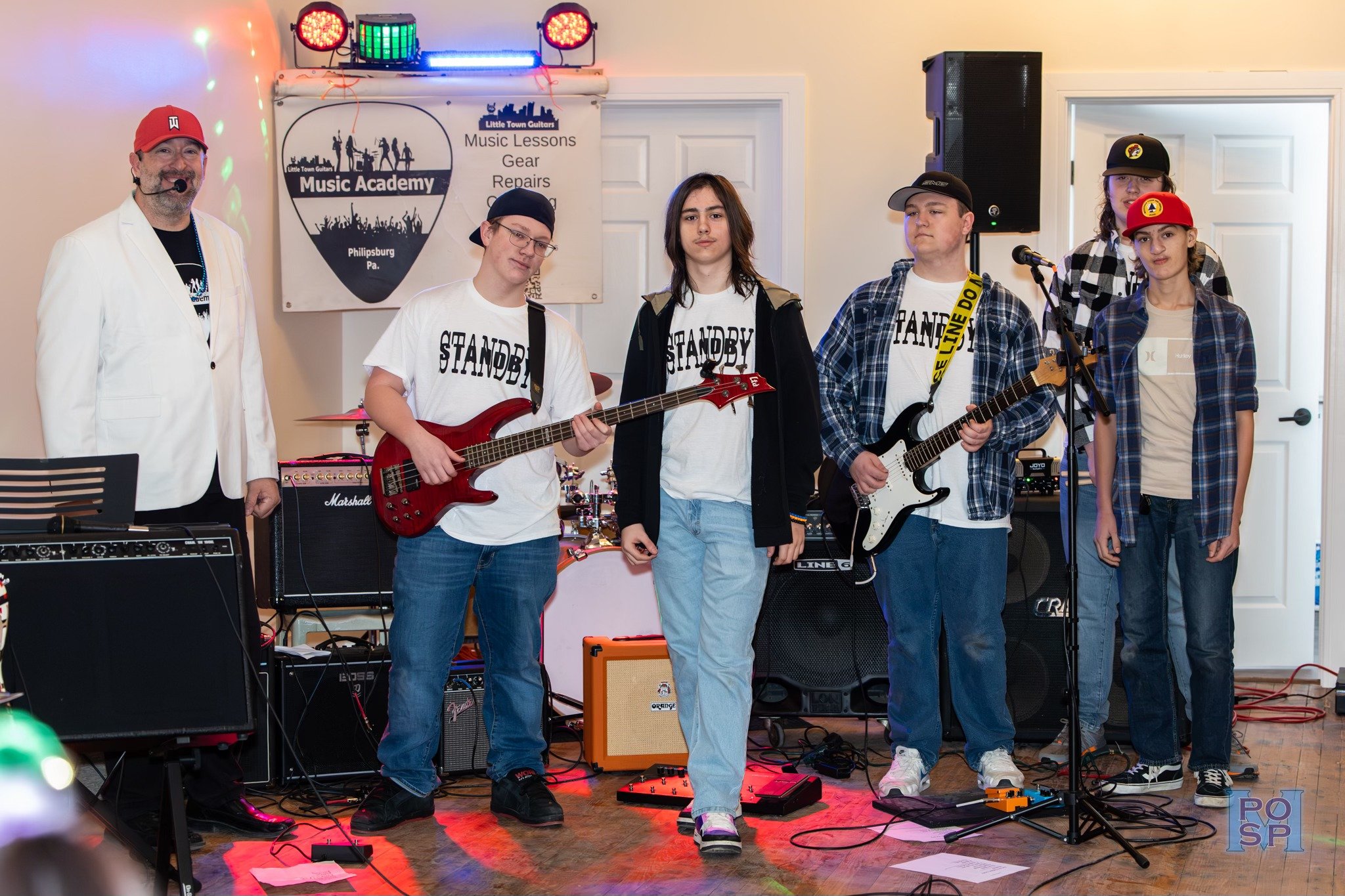 A group of young musicians standing on stage with one adult, possibly a teacher or instructor, during a music lesson or performance at Little Town Guitars Music Academy in Philipsburg, PA. The children are holding guitars and wearing casual attire; s