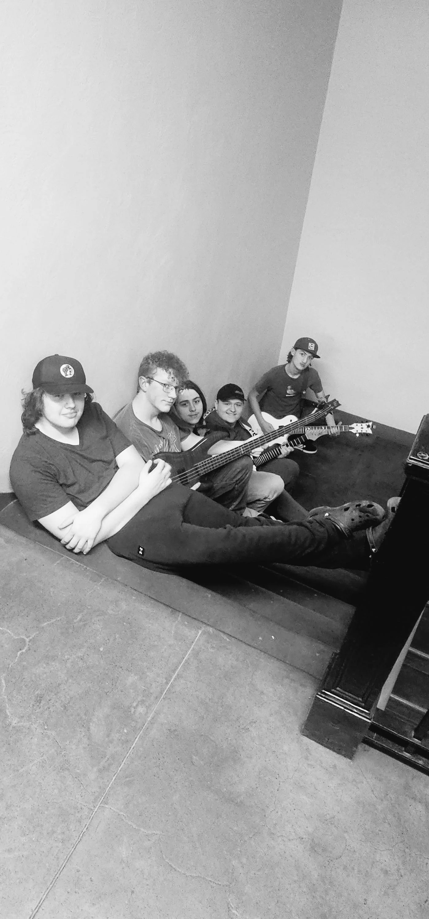 Five teenagers sitting on the floor against a wall, holding musical guitars, with one wearing a baseball cap and sunglasses, in a practice or casual setting.