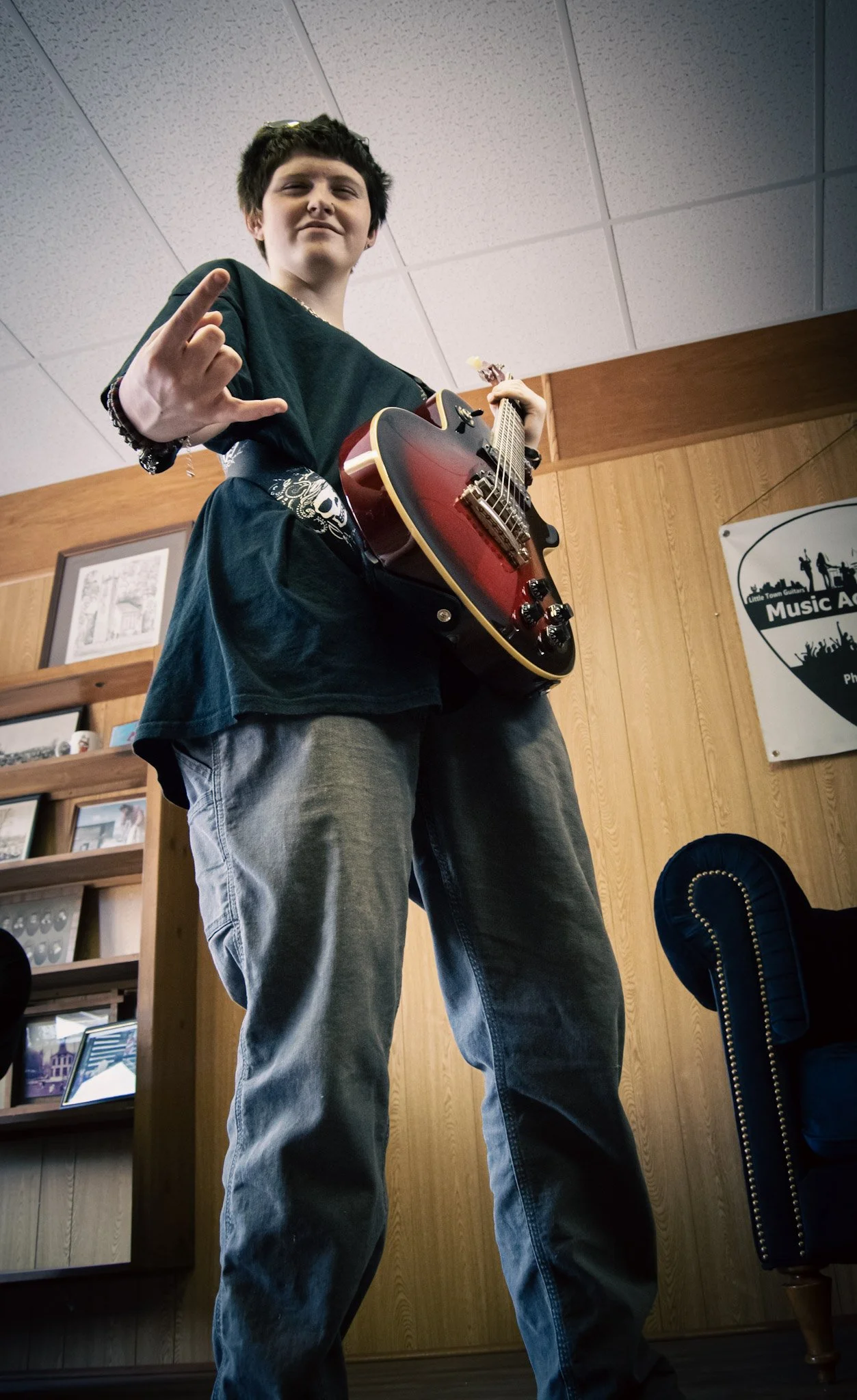 Young person standing indoors, holding a red and black electric guitar, making a rock hand gesture, with wood-paneled walls and a banner in the background.