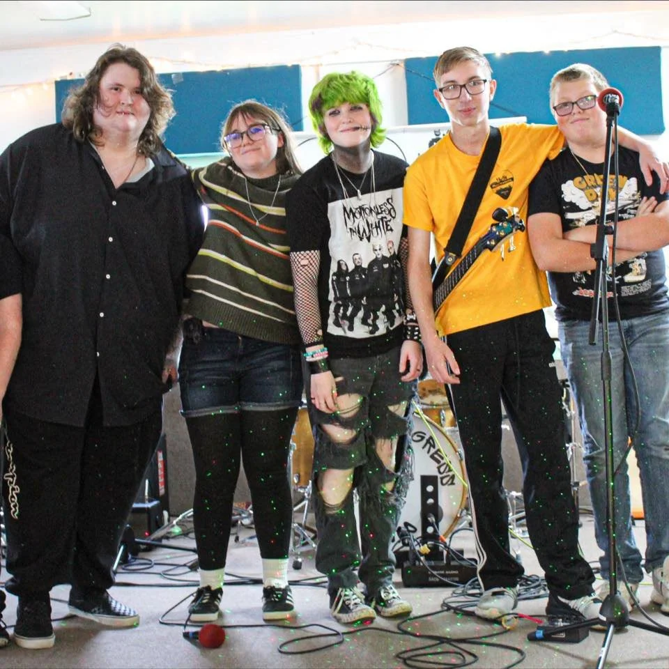 Six young people standing on a stage with musical instruments, wearing casual and punk-style clothing, in front of blue soundproof panels.