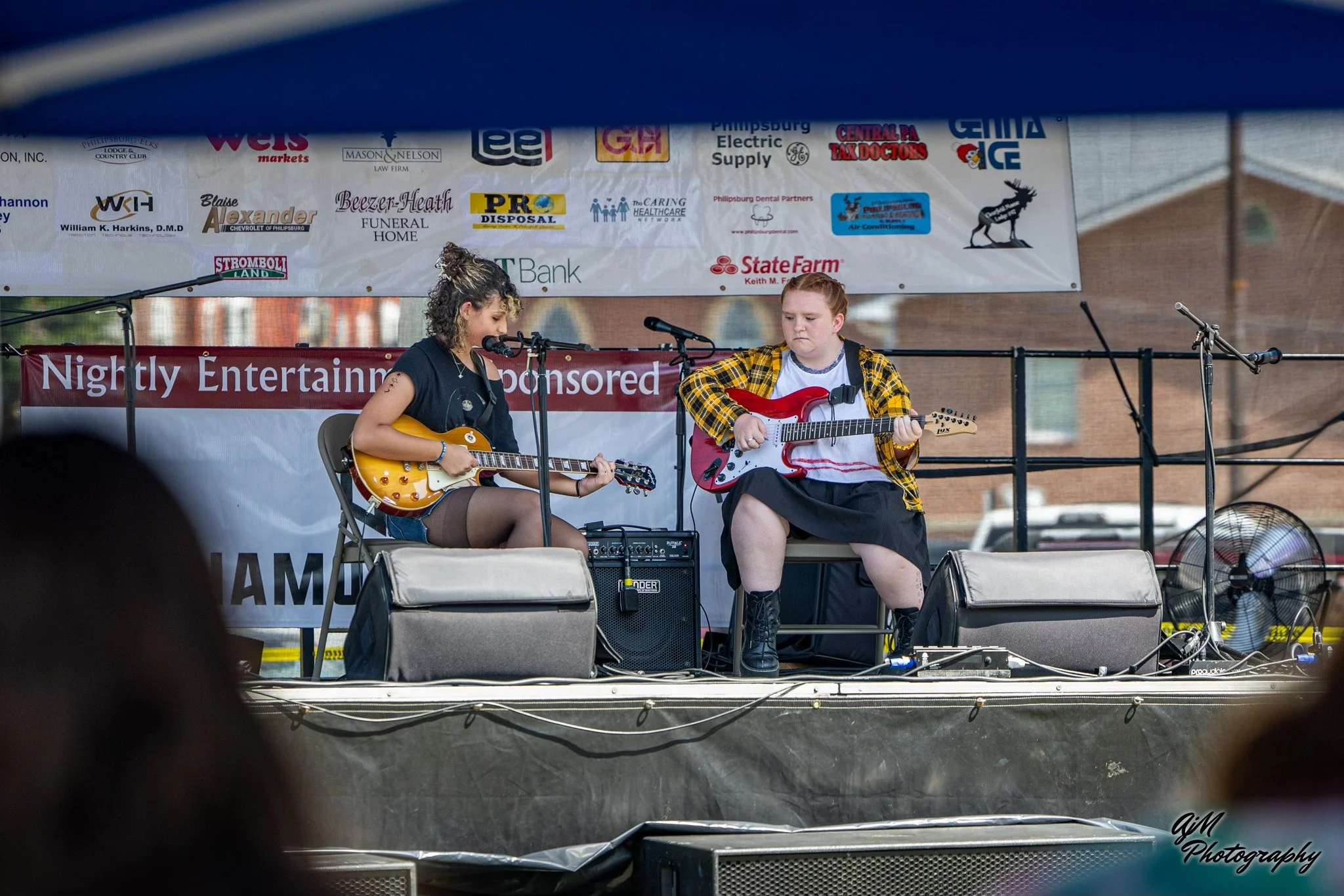 Two young women sitting on a stage playing guitars. One with a yellow guitar and the other with a red guitar. They are performing at an outdoor event with a banner behind them and various sponsor logos above.