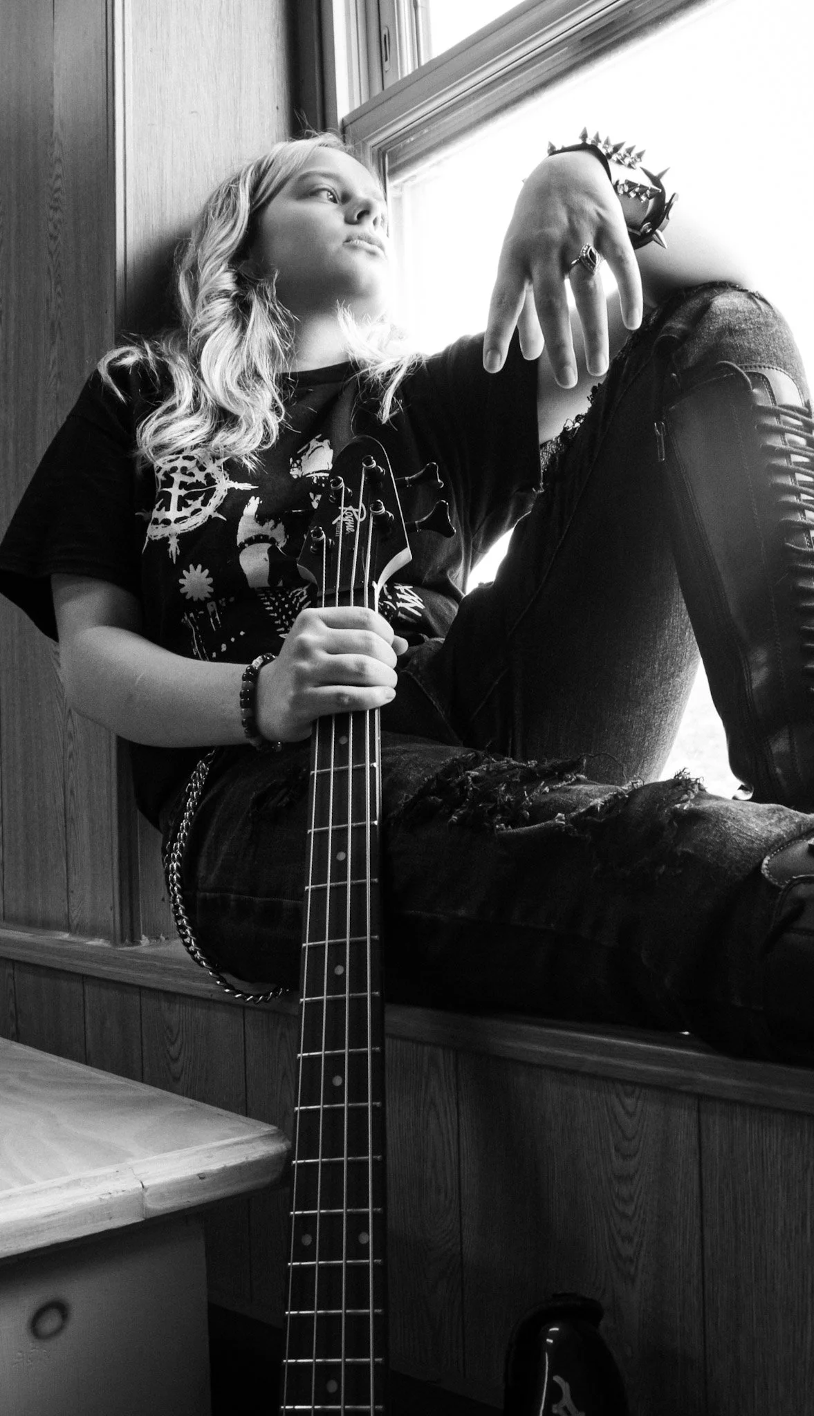 Young woman with long wavy hair, wearing a graphic black T-shirt and ripped jeans, sitting by a window holding a guitar, looking pensively outside in black and white.