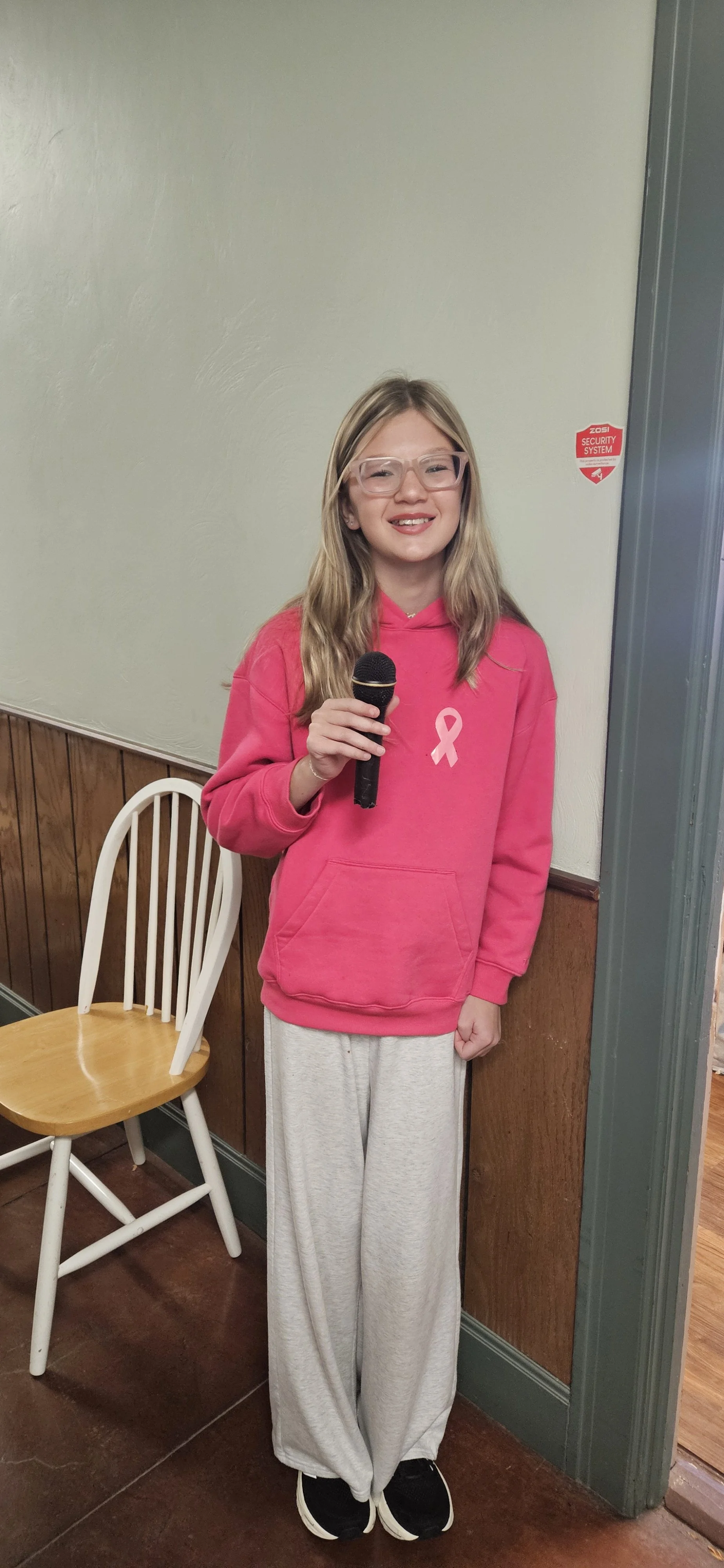 A young woman wearing glasses and a pink hoodie with a breast cancer awareness ribbon stands indoors holding a microphone, smiling at the camera.