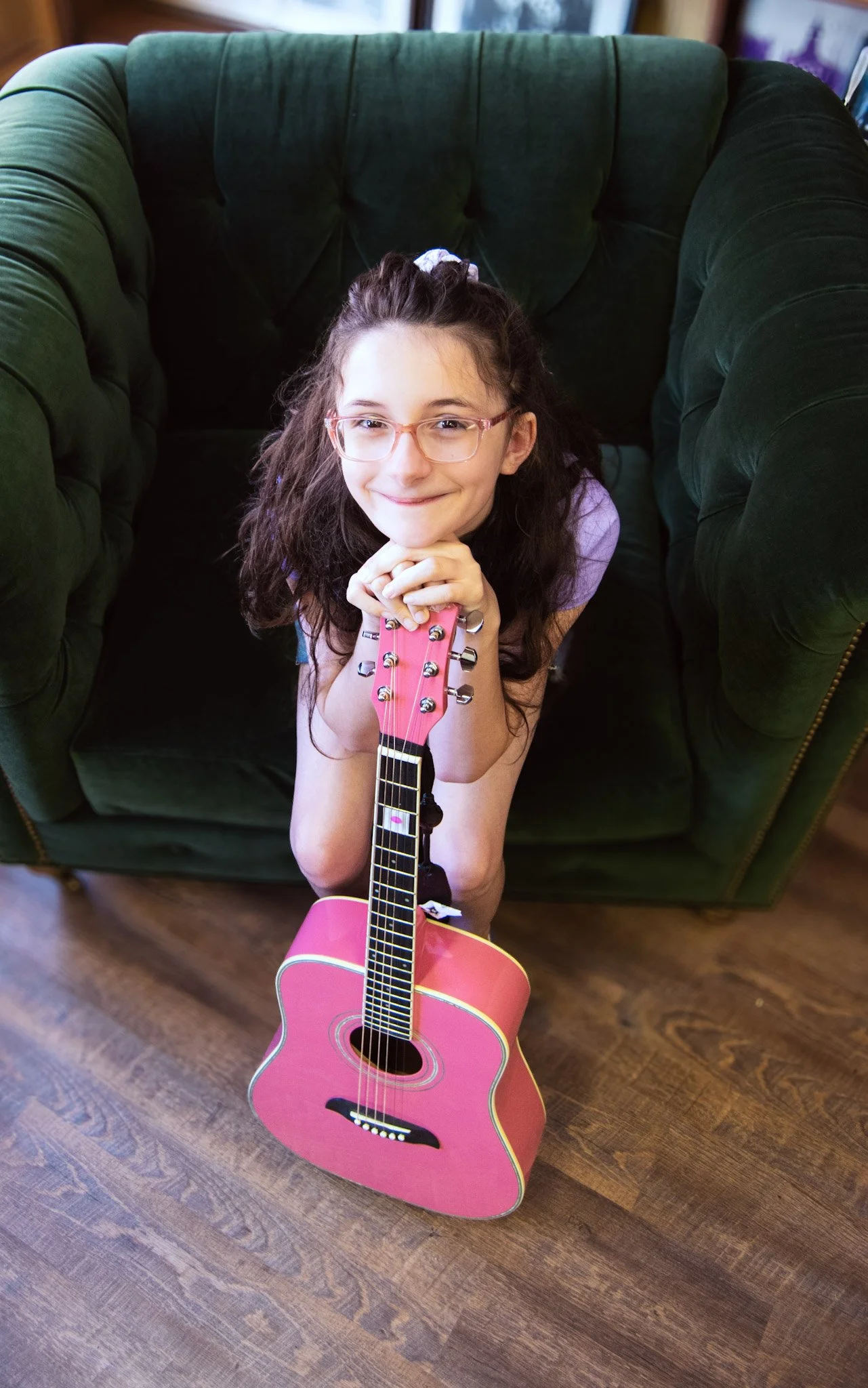 A young girl with brown hair, glasses, and a purple shirt is sitting on her knees on a wooden floor, smiling, and holding a pink acoustic guitar.