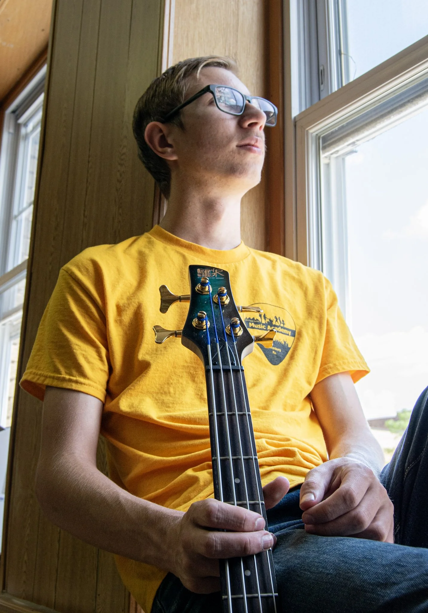 A young man with glasses wearing a yellow t-shirt, sitting indoors near a window, holding a bass guitar.