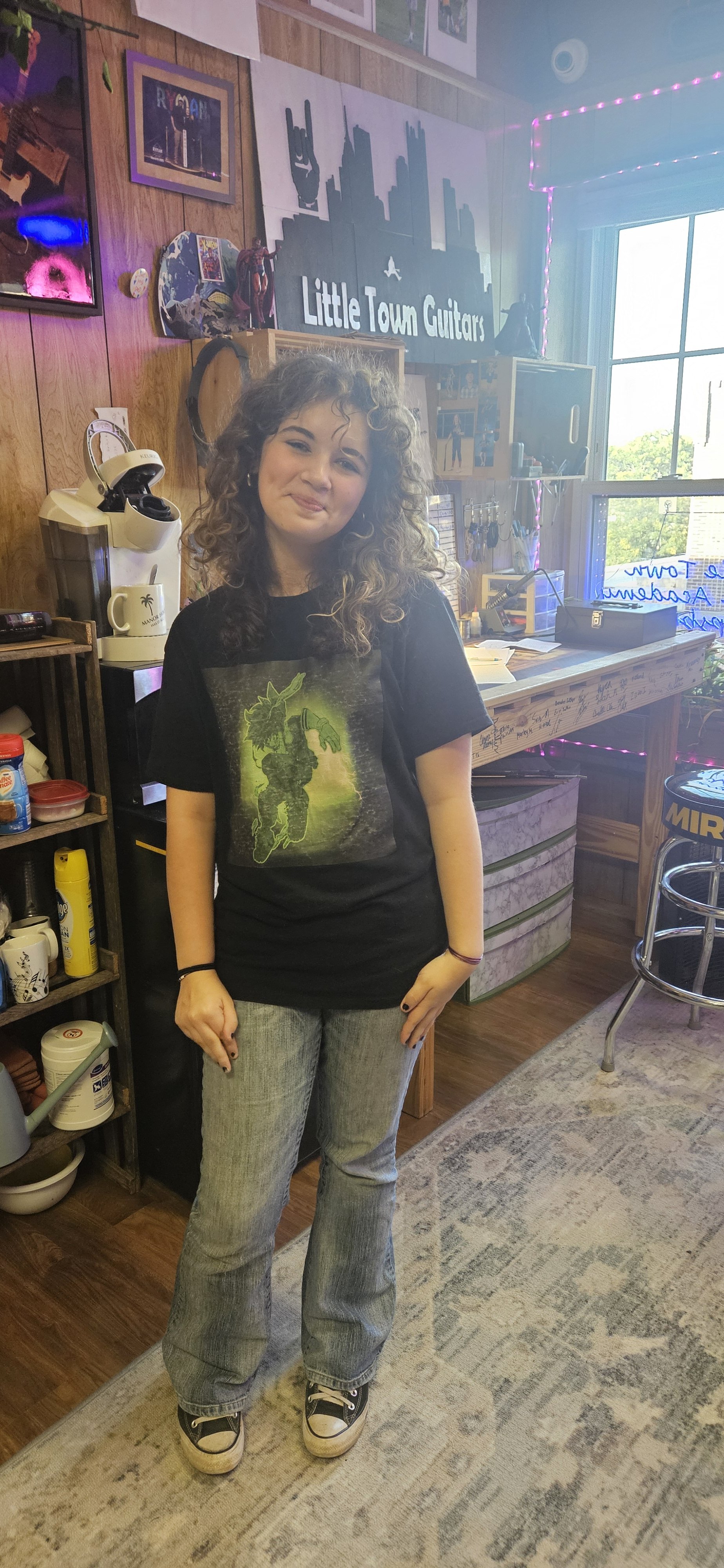 A young woman with curly hair stands in a cozy, decorated room with wooden walls and a window. She wears a black graphic t-shirt, gray jeans, and Converse sneakers. Behind her are shelves with kitchen items and a large sign that reads 'Little Town Gu