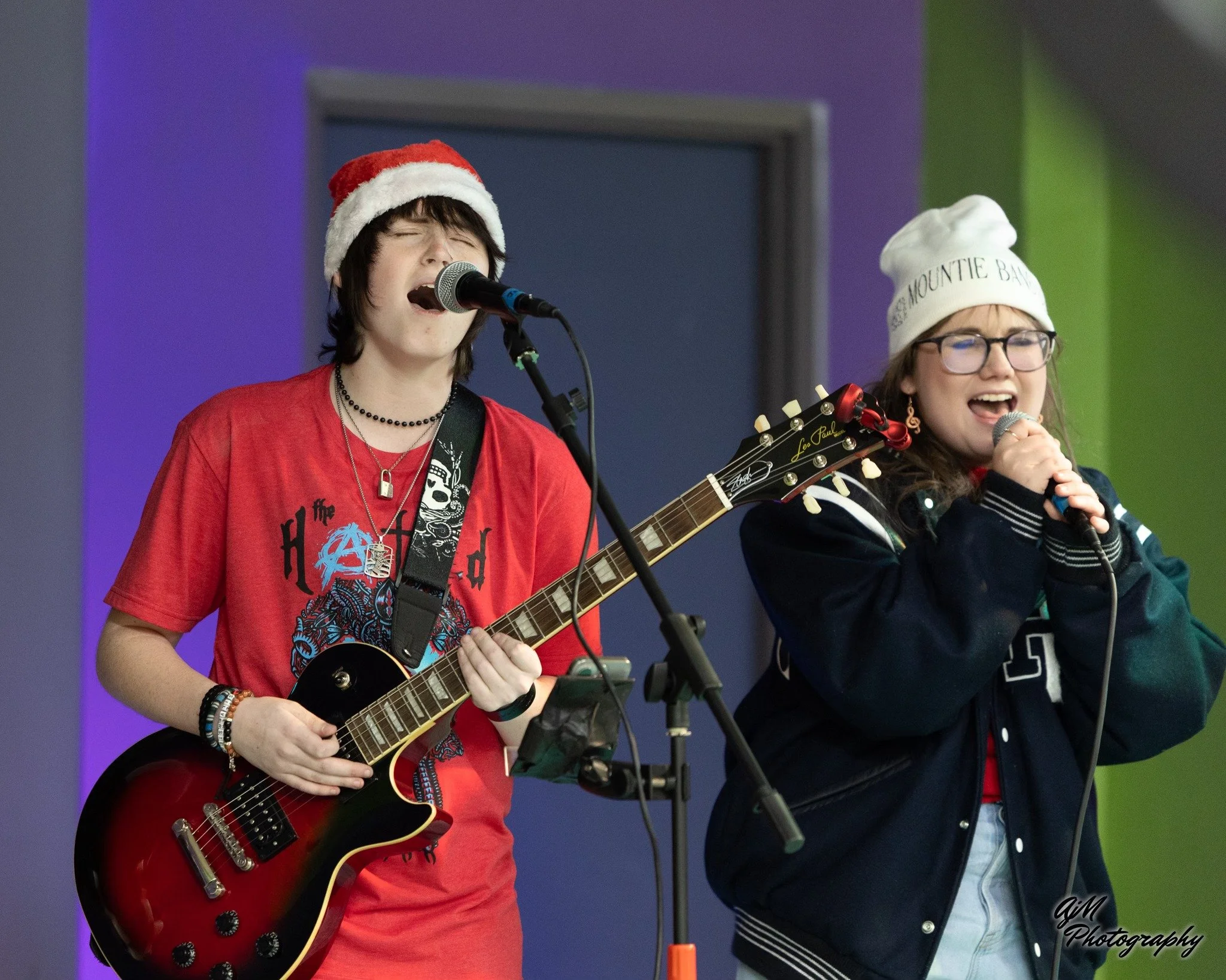 Two teenagers performing music on stage, one playing guitar and singing, wearing a Santa hat, and the other singing into a microphone, wearing a beanie, both with closed eyes and joyful expressions.