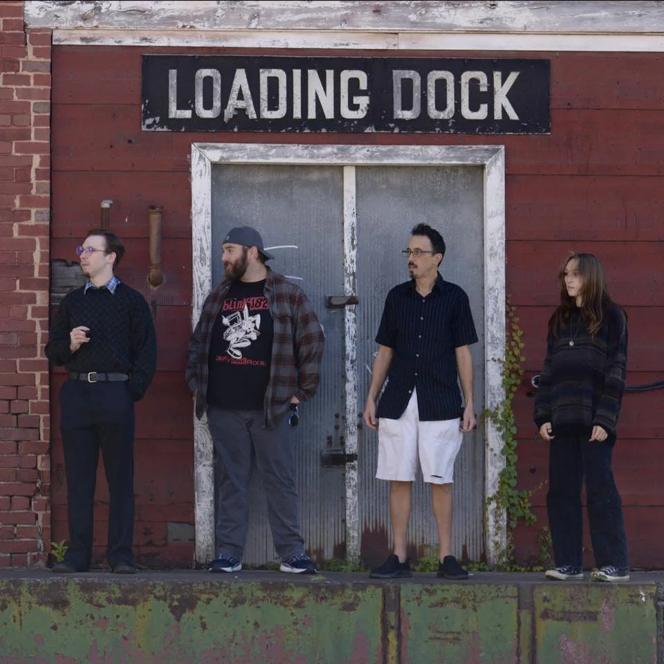 Four people standing in front of a weathered loading dock door on a brick and wood wall.