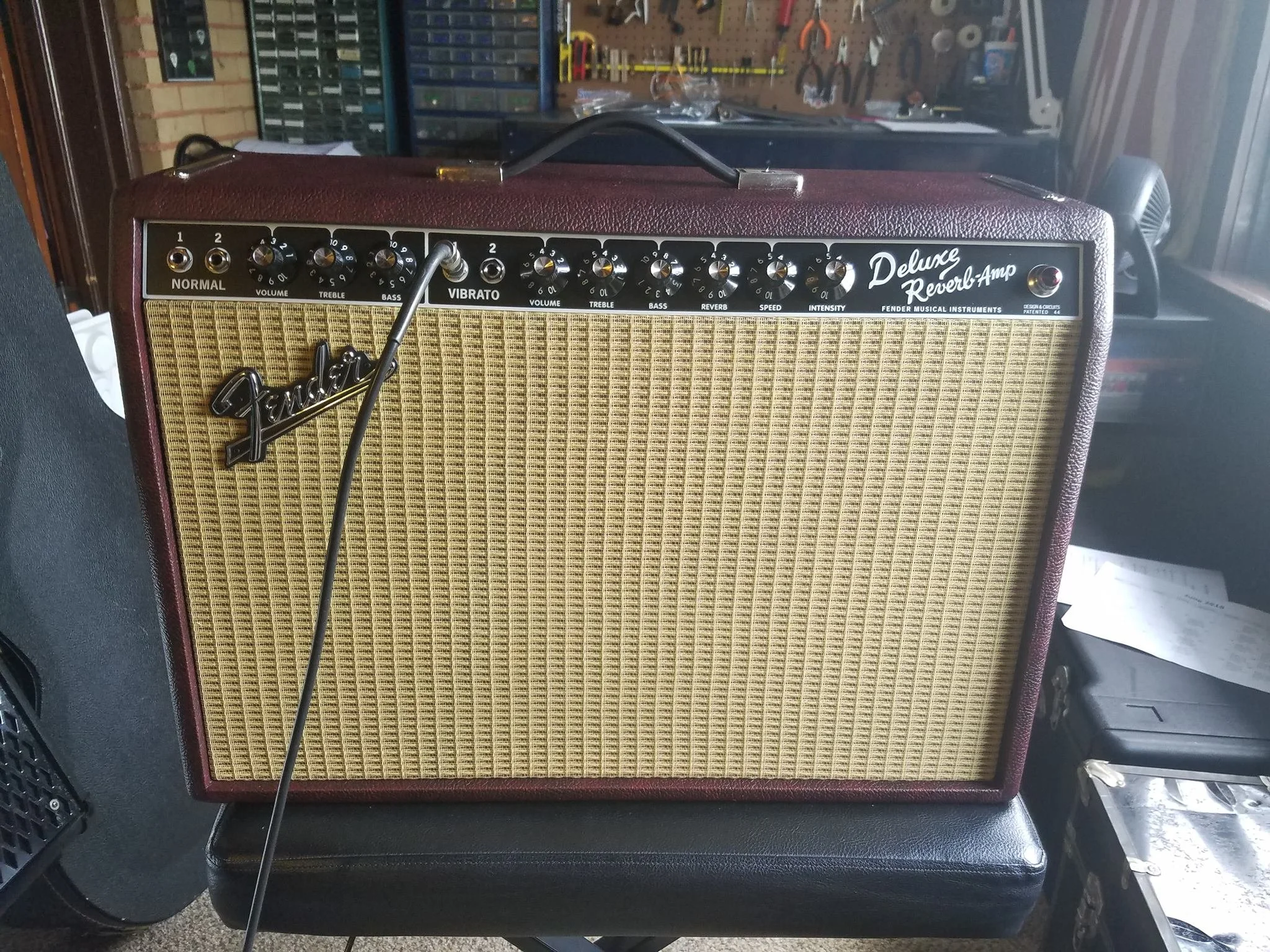A Fender Deluxe Reverb guitar amplifier with a purple and beige exterior, control knobs on top, and a black handle, placed on a black surface in a room with musical equipment and tools in the background.