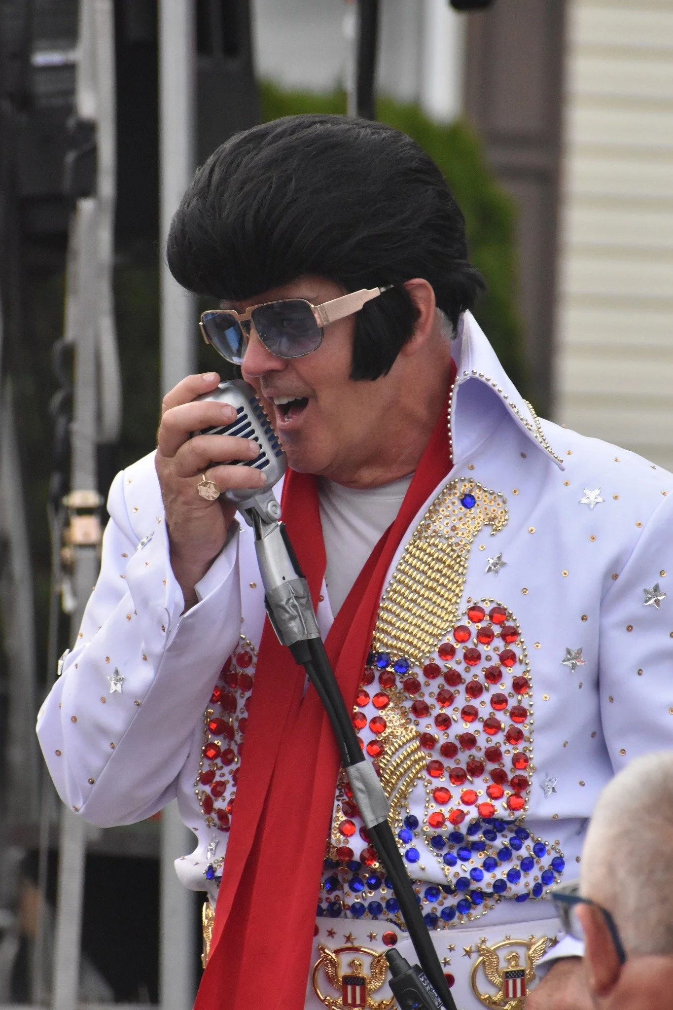 Elvis impersonator singing into a microphone outdoors, wearing a glittery white jumpsuit with a red scarf and large sunglasses.