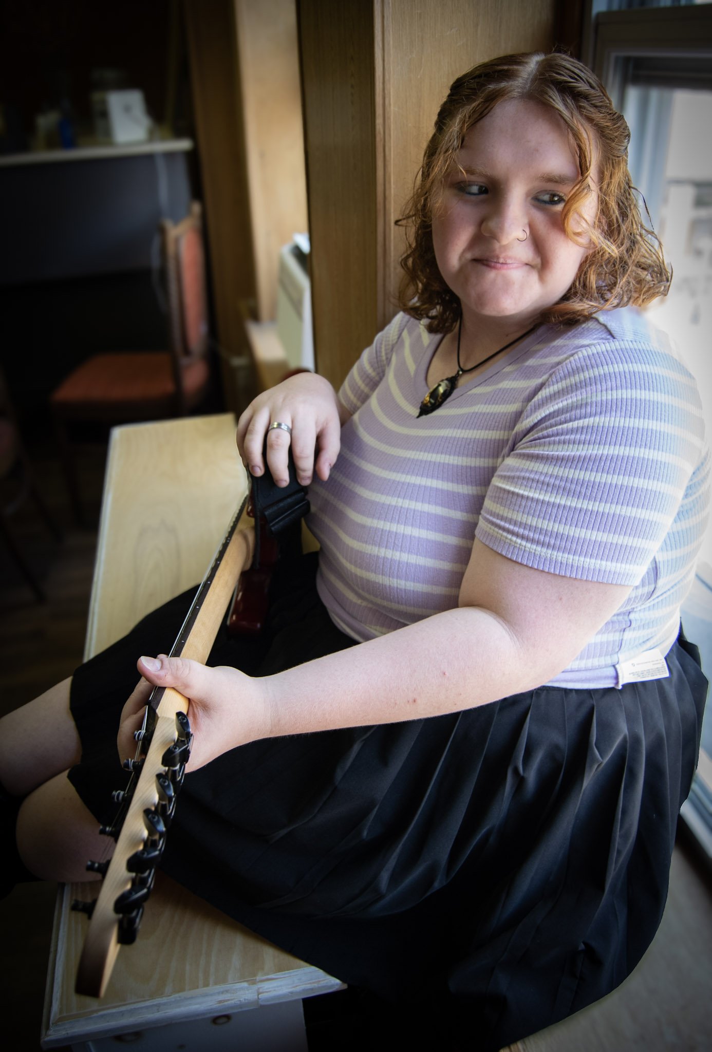 A young woman with curly red hair and a nose piercing, sitting on a wooden chair, holding a guitar on her lap, in a cozy indoor setting.