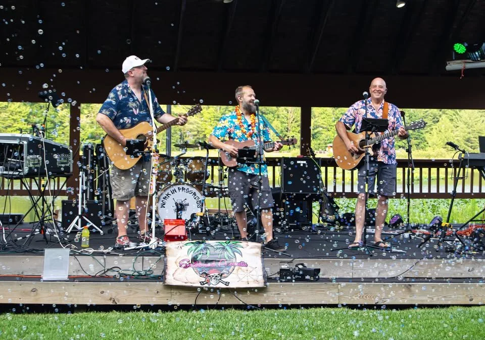 Four male musicians wearing Hawaiian shirts and shorts performing on an outdoor stage with instruments, surrounded by confetti and greenery in the background.