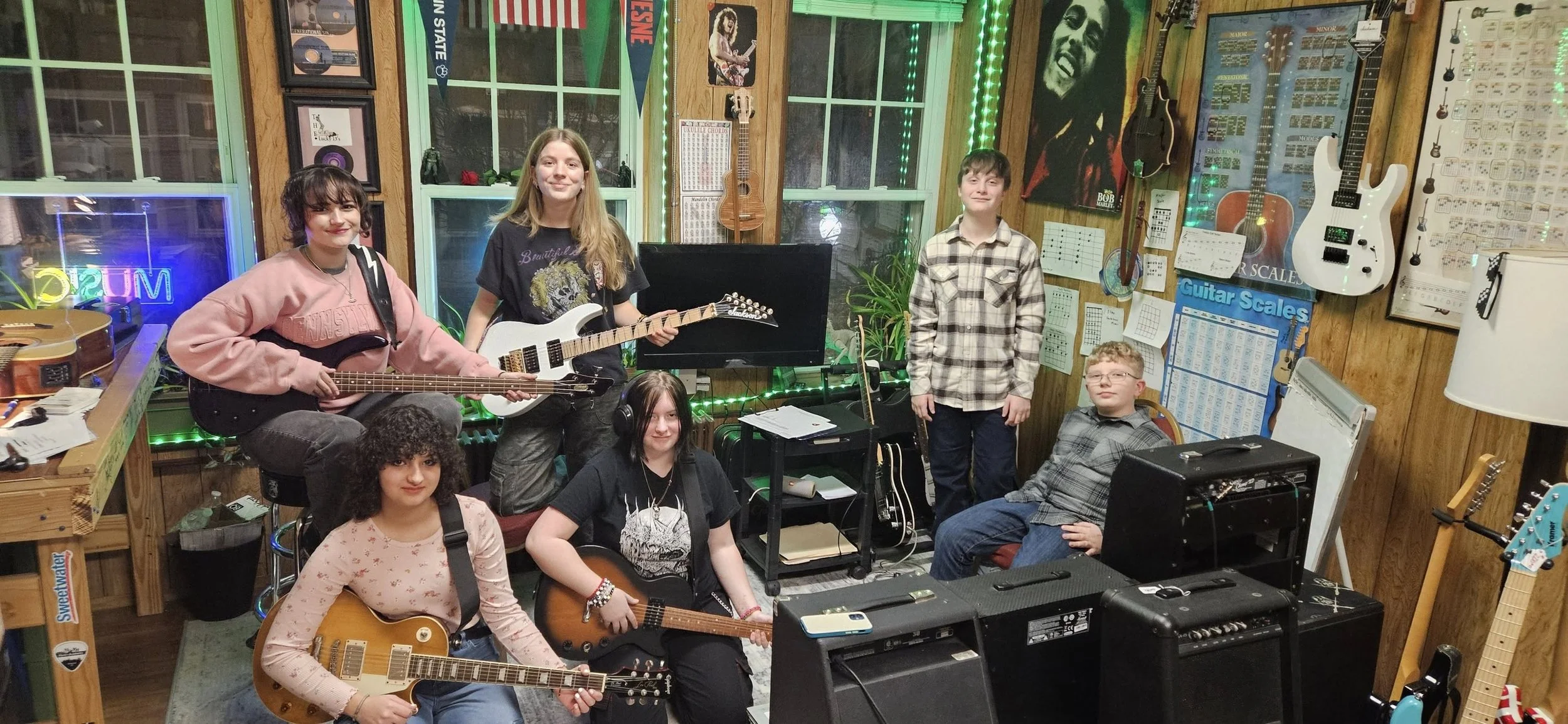 A group of seven young people in a music studio. Some are sitting and others are standing, holding guitars. The room is decorated with music posters, guitars on the wall, and a neon sign that says "JUMP." There is music equipment, a black music stand