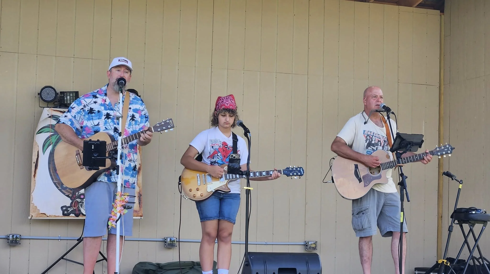 Three musicians playing guitars and singing on stage, with a beige wooden background. The man on the left wears a colorful Hawaiian shirt, shorts, and a hat. The girl in the middle wears a red bandana, a white t-shirt with a comic character, and deni
