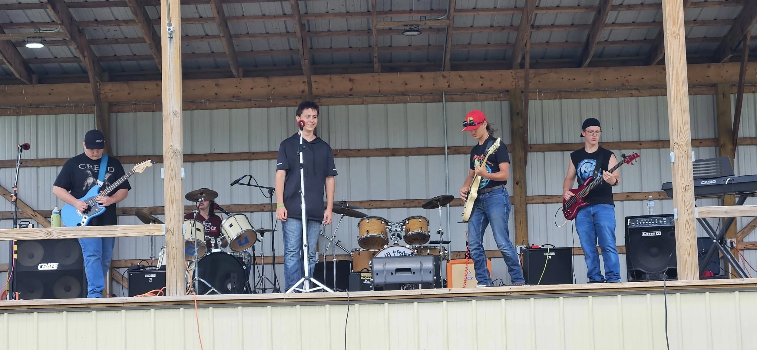 A band of five young musicians performing on a wooden stage in a barn-like structure. They are playing various instruments including electric guitars, a bass, and drums, with one person singing into a microphone.