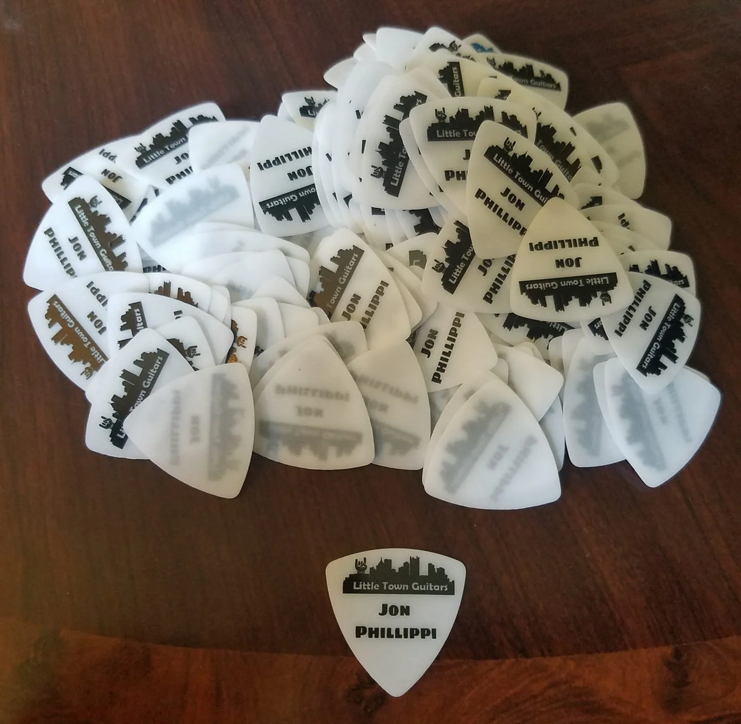 Pile of white guitar picks with black text and logo, some picks are scattered in front, on a wooden surface.