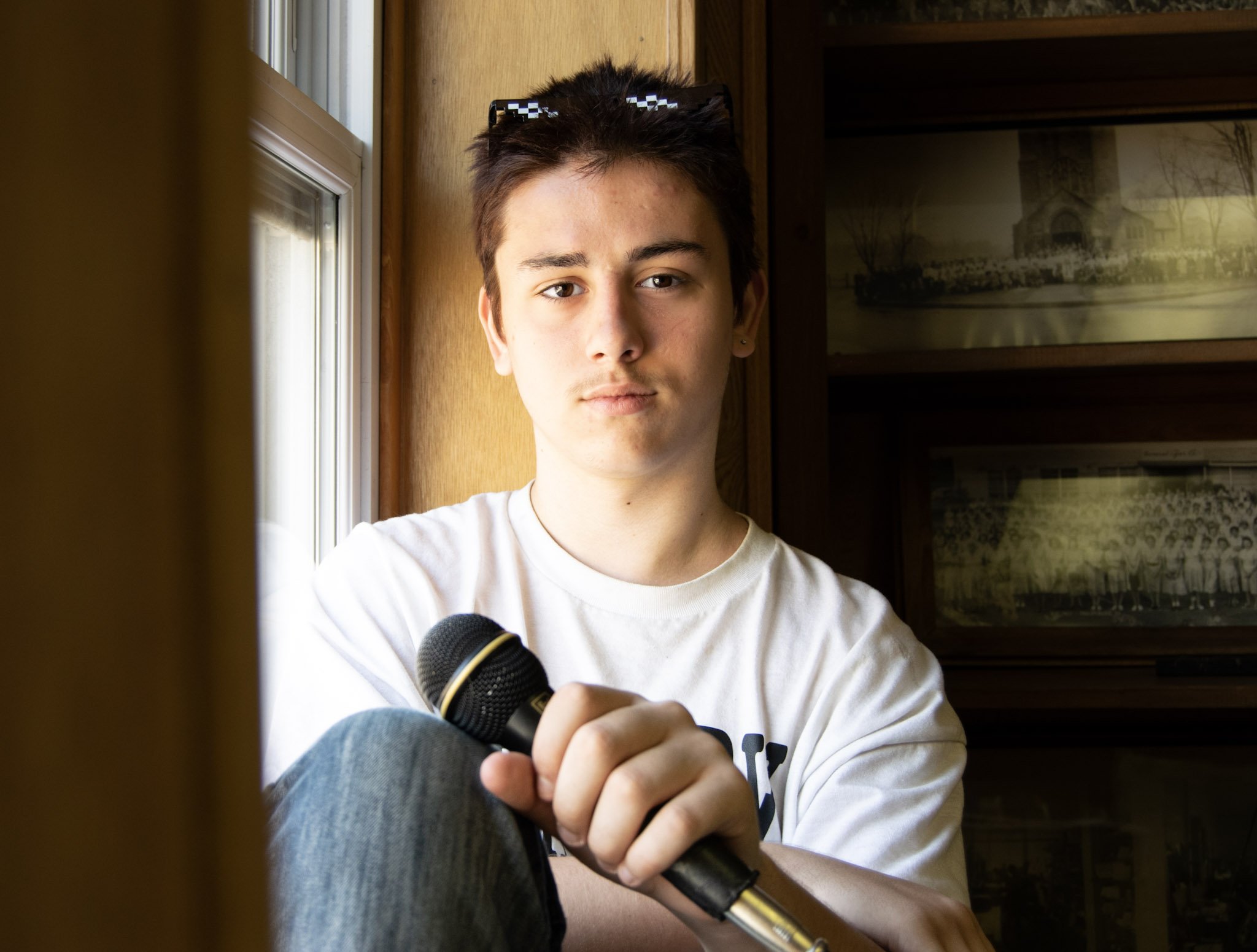 Young person with short dark hair sitting by a window, holding a microphone, wearing a white T-shirt, with framed pictures in the background.