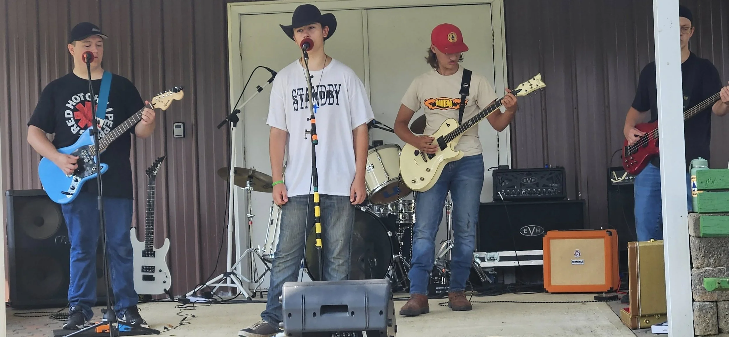 A group of five young men performing on stage, playing guitars and singing, with a drum set in the background and various amplifiers and equipment around them.