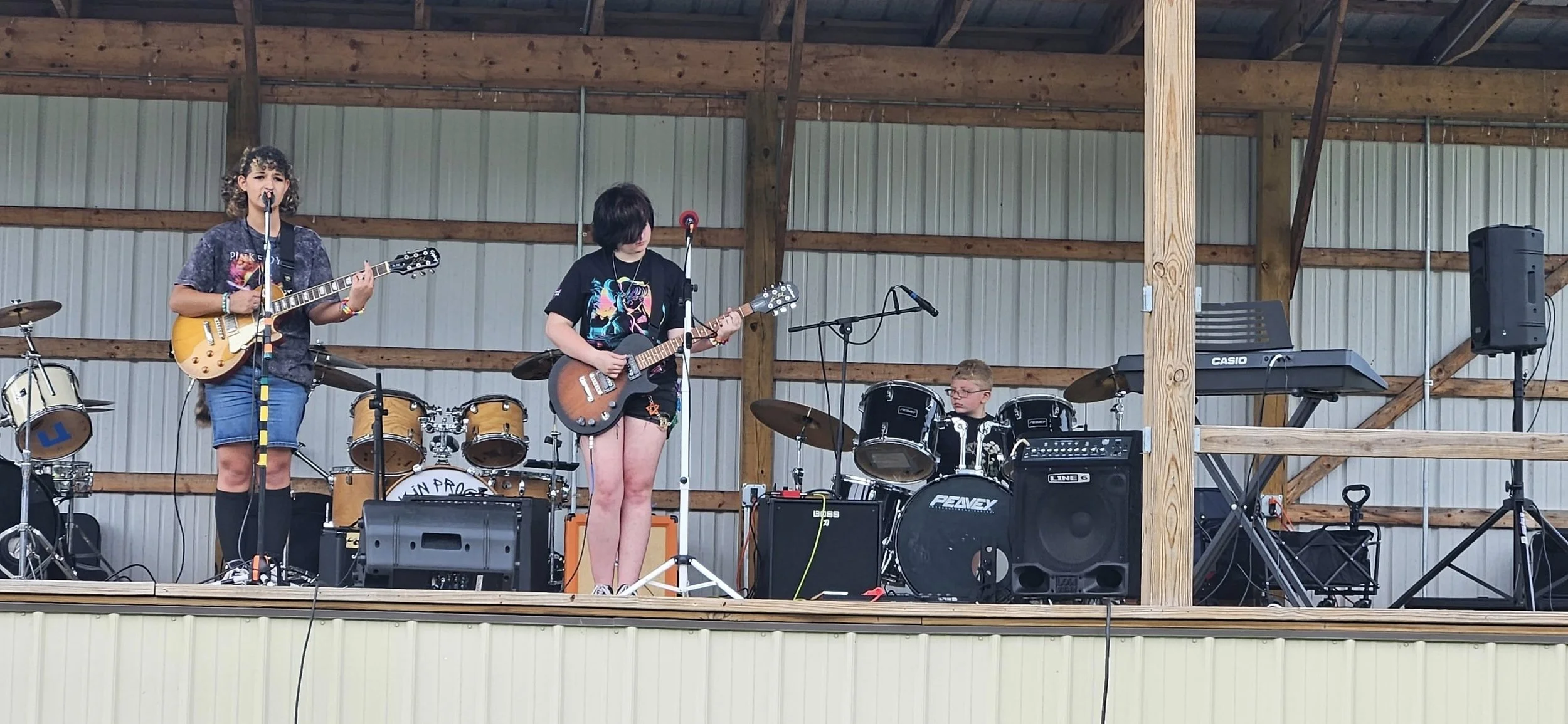 Three young musicians performing on a wooden stage inside a barn-like structure. The girl on the left is singing into a microphone and playing an electric guitar, wearing a graphic T-shirt, shorts, and knee-high socks. The girl in the middle is also 
