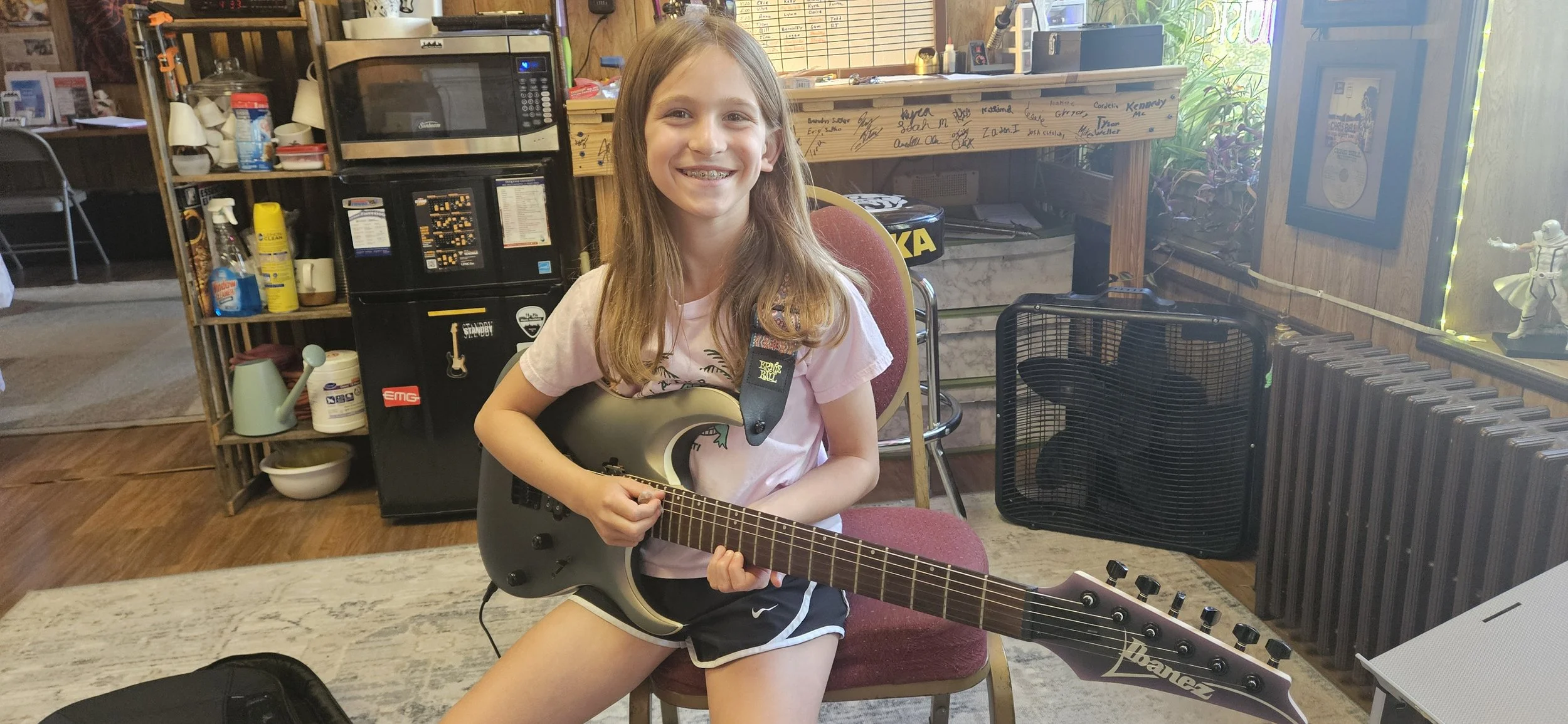 A young girl with long blonde hair and braces, sitting on a red chair, smiling and holding a black electric guitar in a casual indoor setting with wood-paneled walls, a window, and various furniture and decorations.