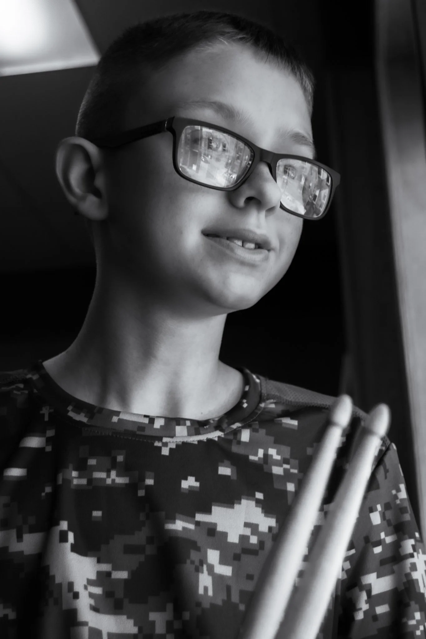 A black and white photo of a young boy wearing glasses, a camo-patterned shirt, and holding drumsticks, looking out a window.