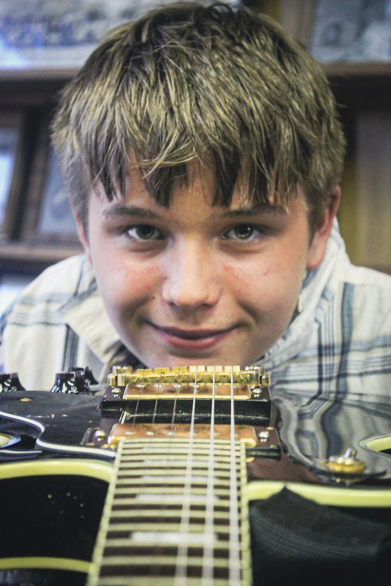 Young boy leaning over an electric guitar, smiling, with shelves of records in the background.