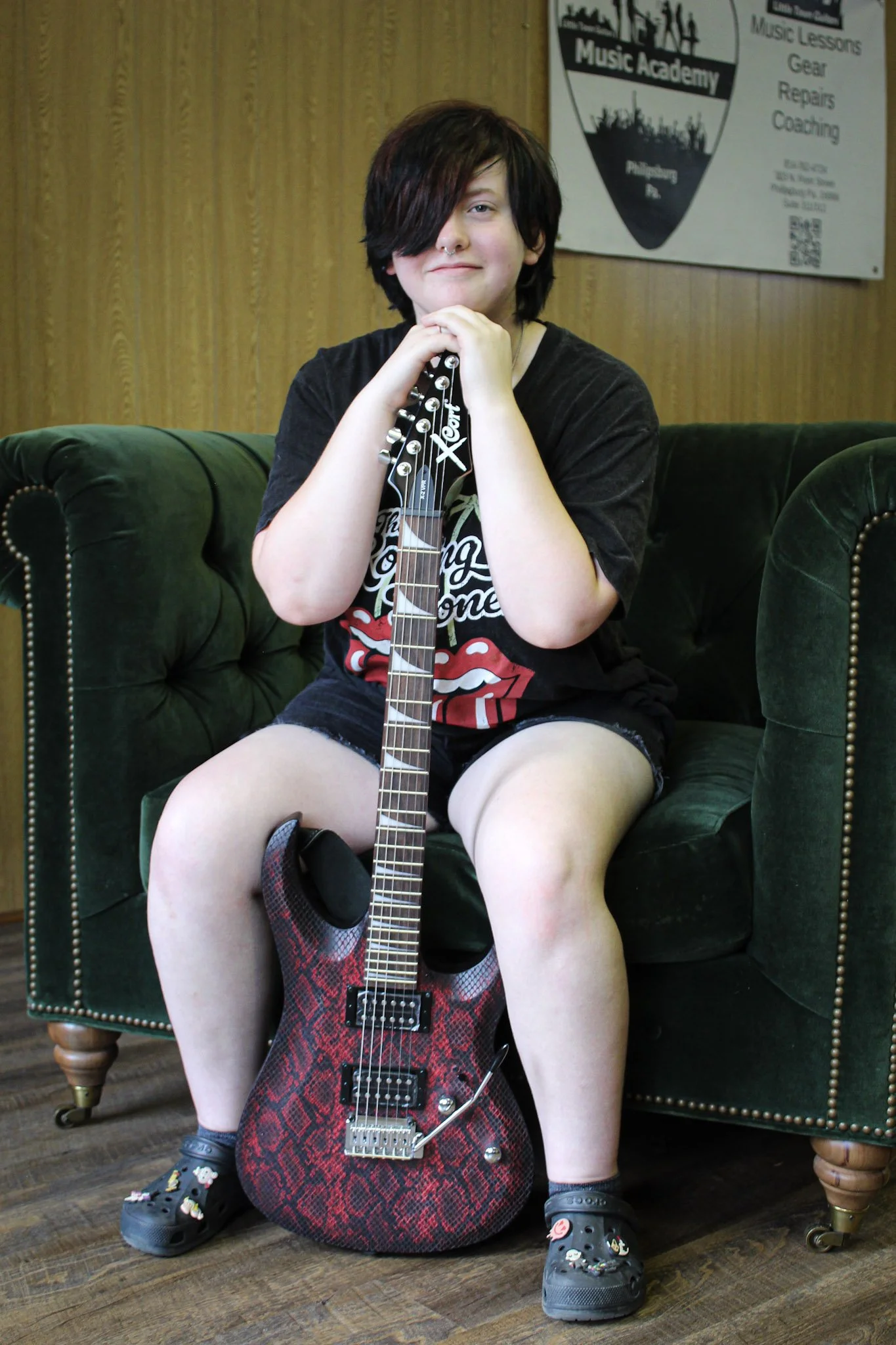 A young person with dark hair and a nose piercing sitting on a green velvet couch, holding an electric guitar with a red and black snakeskin pattern, inside a music academy.