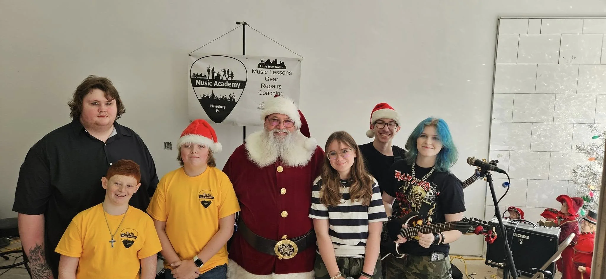 Group of six young people and Santa Claus standing indoors. Some are wearing Santa hats, one holds a guitar. Christmas decorations and a sign for a music academy are in the background.