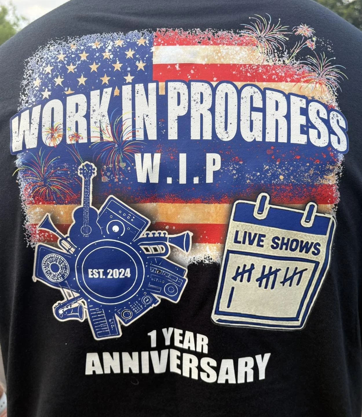 A black T-shirt with patriotic and musical design celebrating one year anniversary of a barbecue work-in-progress event, established in 2024, featuring fireworks, an American flag, musical instruments, speakers, a turntable, and a scoreboard with tally marks.