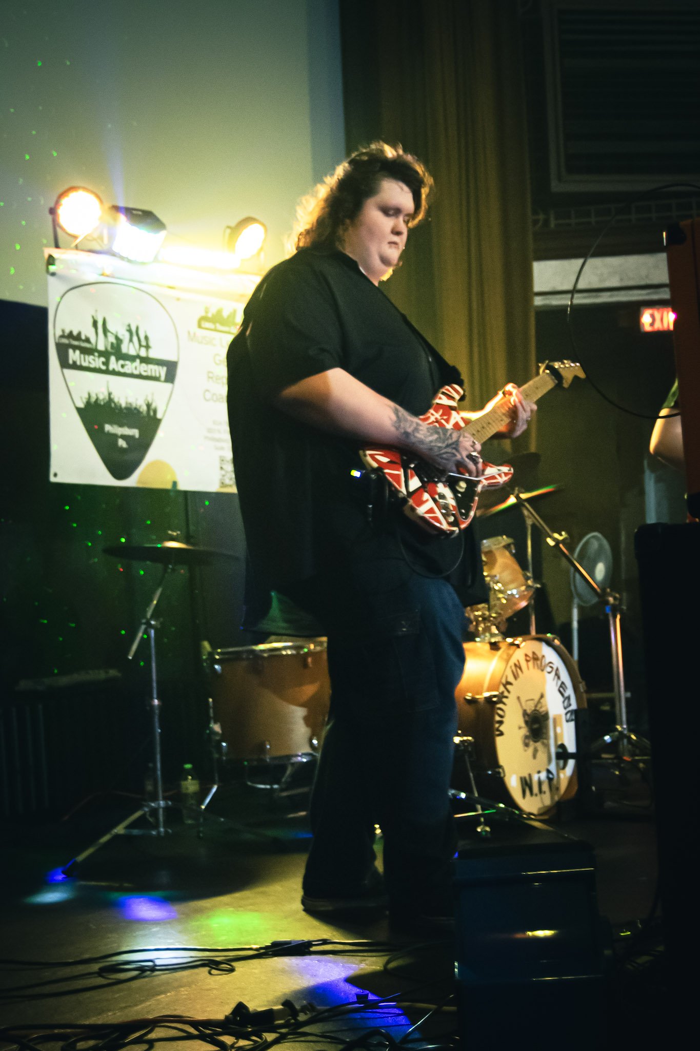 A woman playing an electric guitar on stage at a music performance with a drum set and a sign for Music Academy in the background.