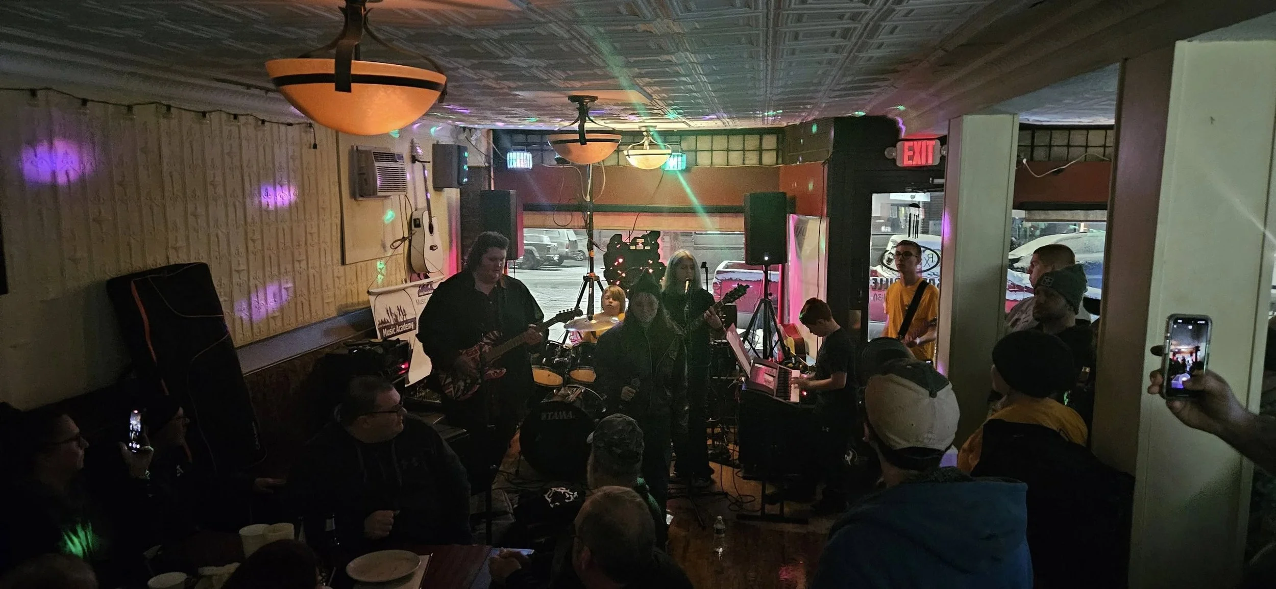 Live music performance at a small indoor venue, with a band playing guitars and a keyboard to an audience, colorful stage lighting, and a view of parked cars outside through the window.