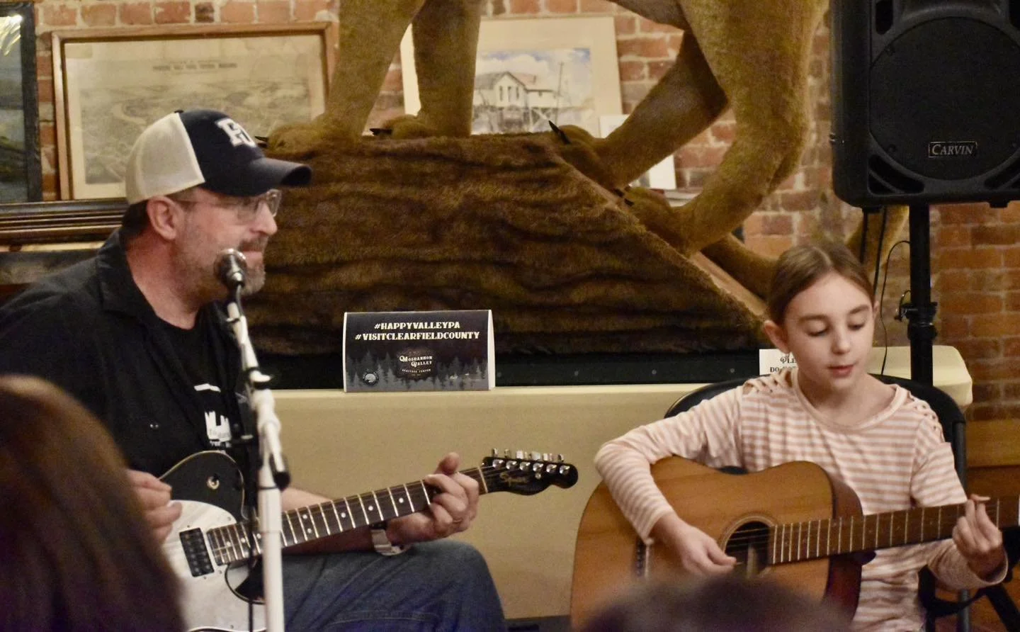 A man with glasses, a beard, and a baseball cap plays an acoustic guitar, while a young girl with brown hair and a striped shirt also plays an acoustic guitar. They are in an indoor setting with exposed brick walls and framed pictures, and there is a