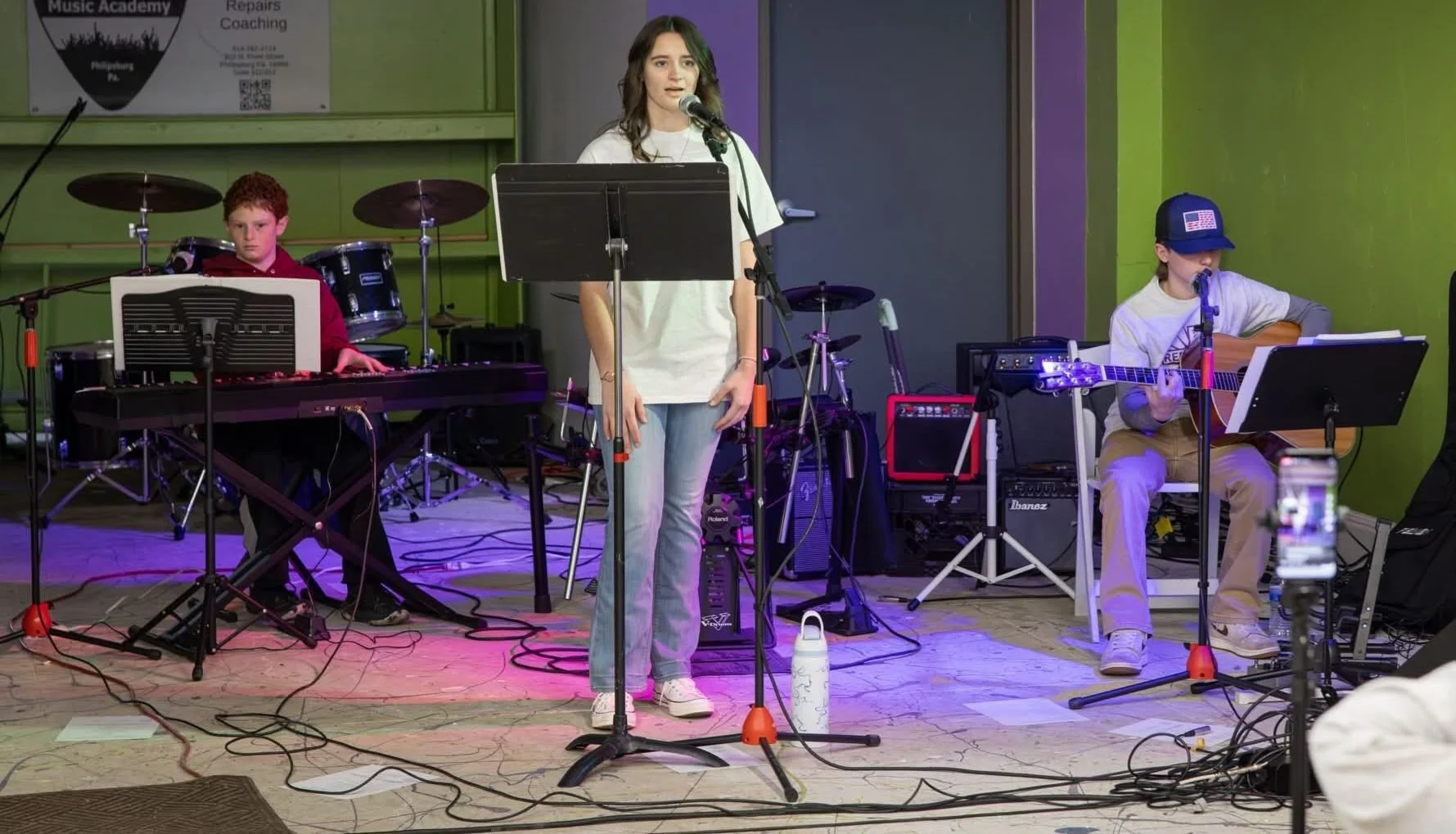 A girl singing into a microphone while two boys play instruments on stage with green walls and musical equipment.