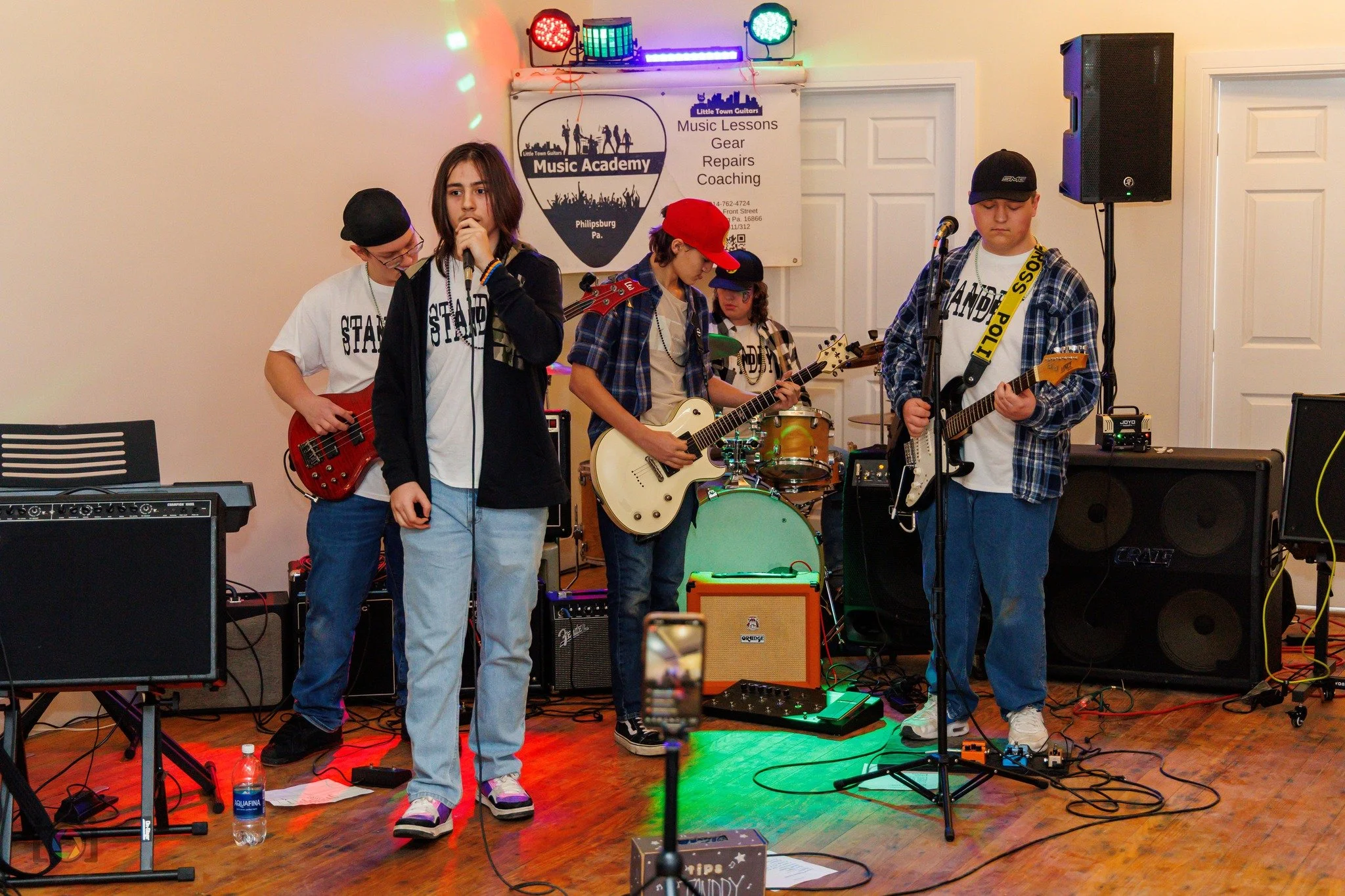 Young children performing a band on stage with musical instruments and equipment, in a room with a sign for a music academy in the background.
