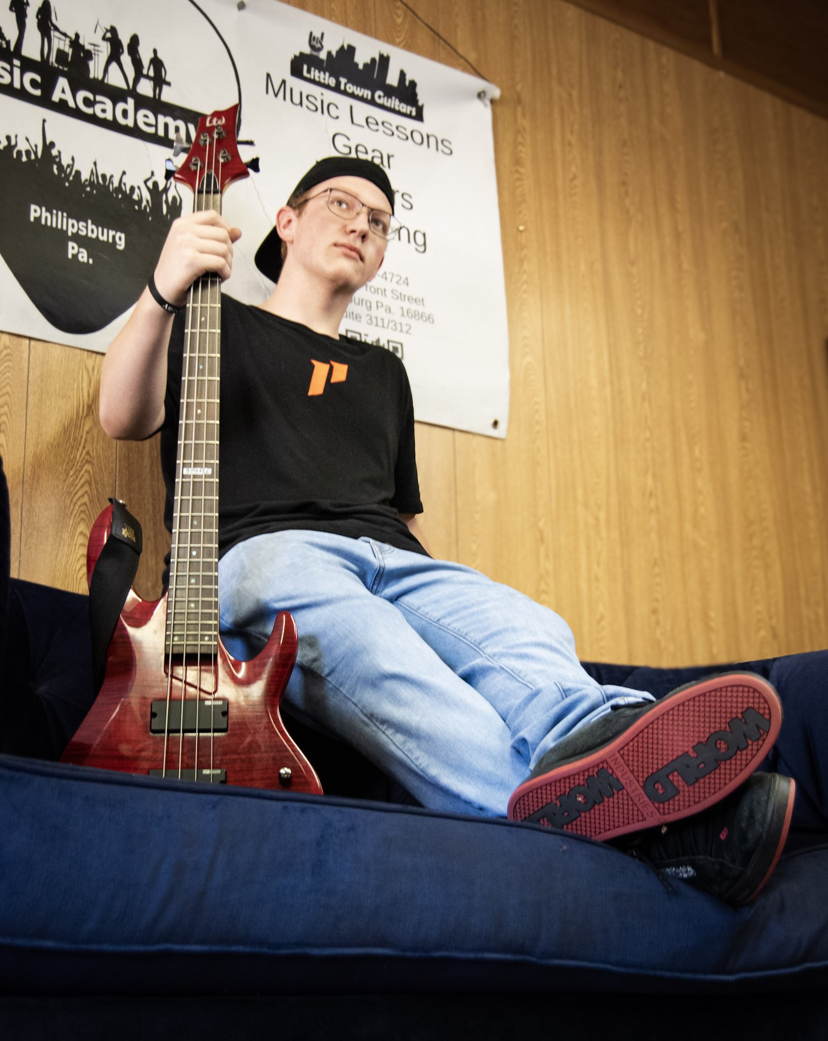 Young man sitting on a couch holding a red bass guitar, wearing a black T-shirt, blue jeans, and black sneakers with red soles, in a music lesson room with wooden panel walls and a promotional banner in the background.